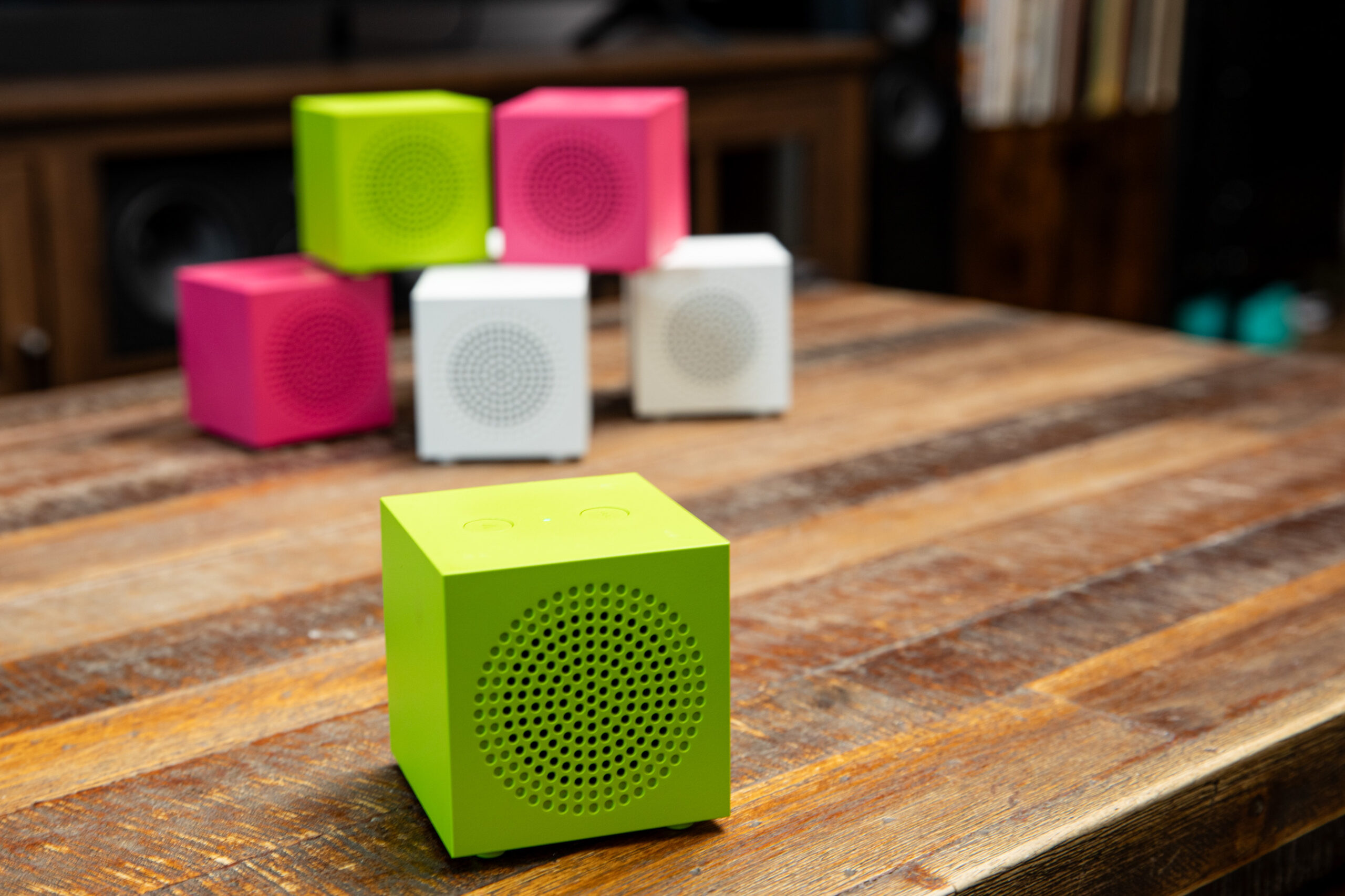 A green Ikea Kallsup Bluetooth speaker on a wooden coffee table in front of a stack of more green, pink, and white Kallsup speakers.