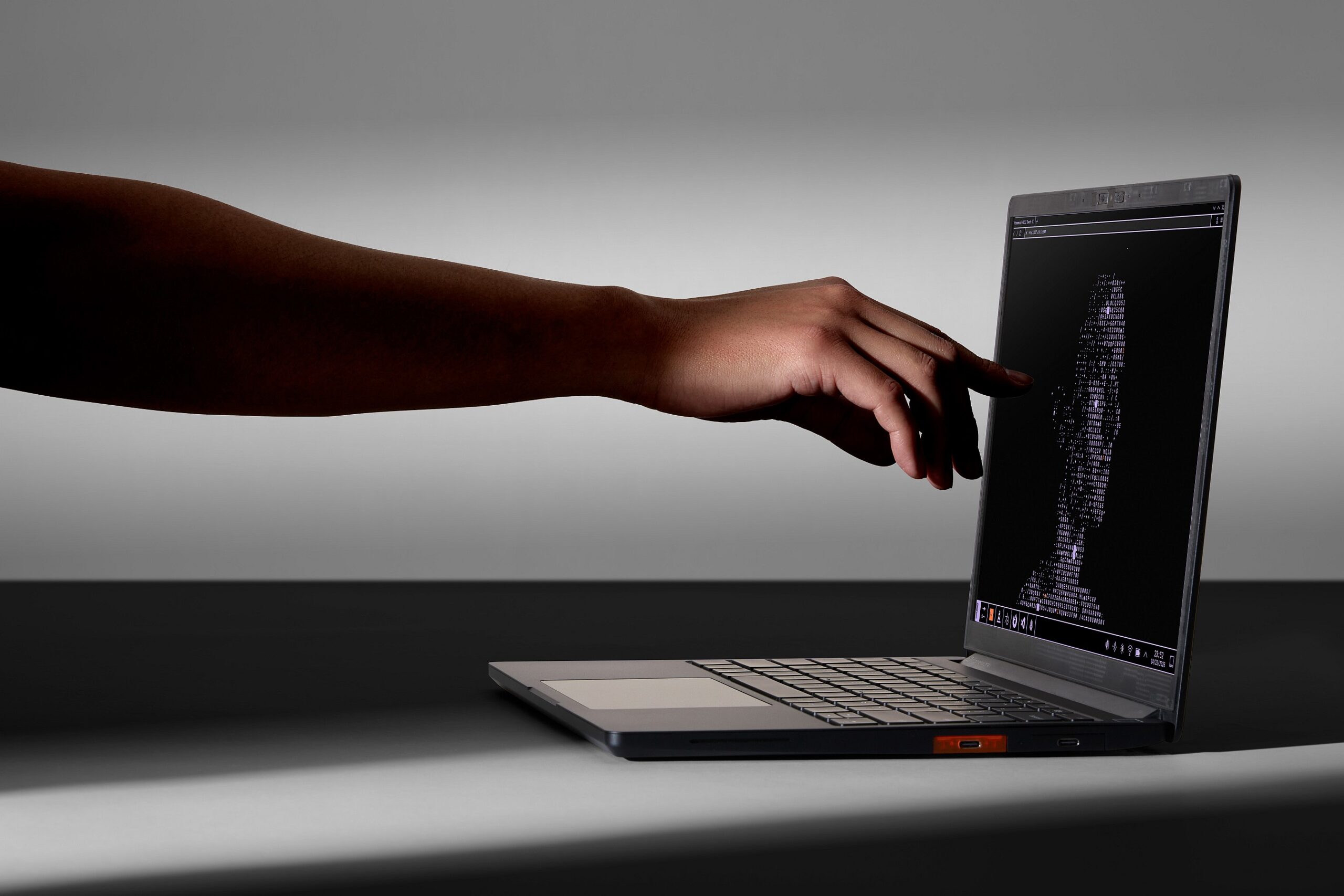 A hand and arm, in shadow, reaches out to touch the screen of the Framework Laptop 13 Pro, a shaft of light illuminating the hand