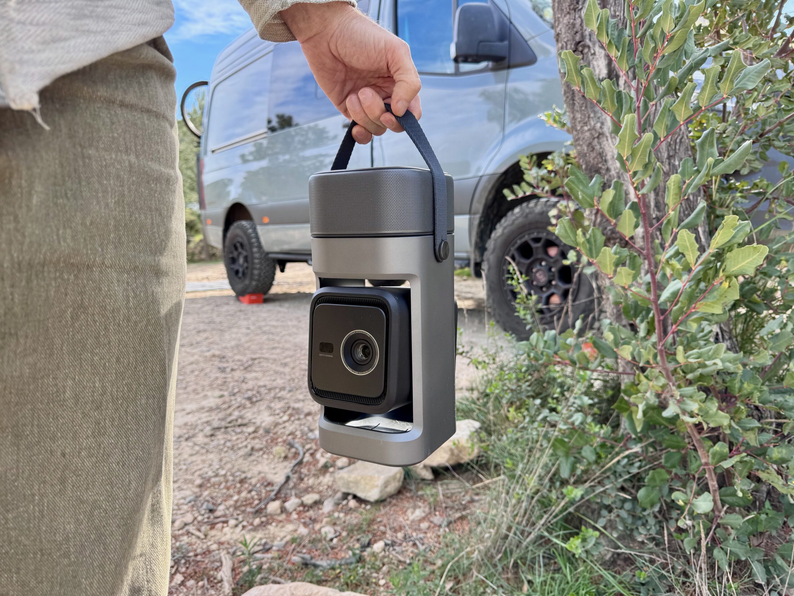 the Nebula P1 is held by its handle with the projector lens exposed in front of a van parked in front of a bush and gravel road.