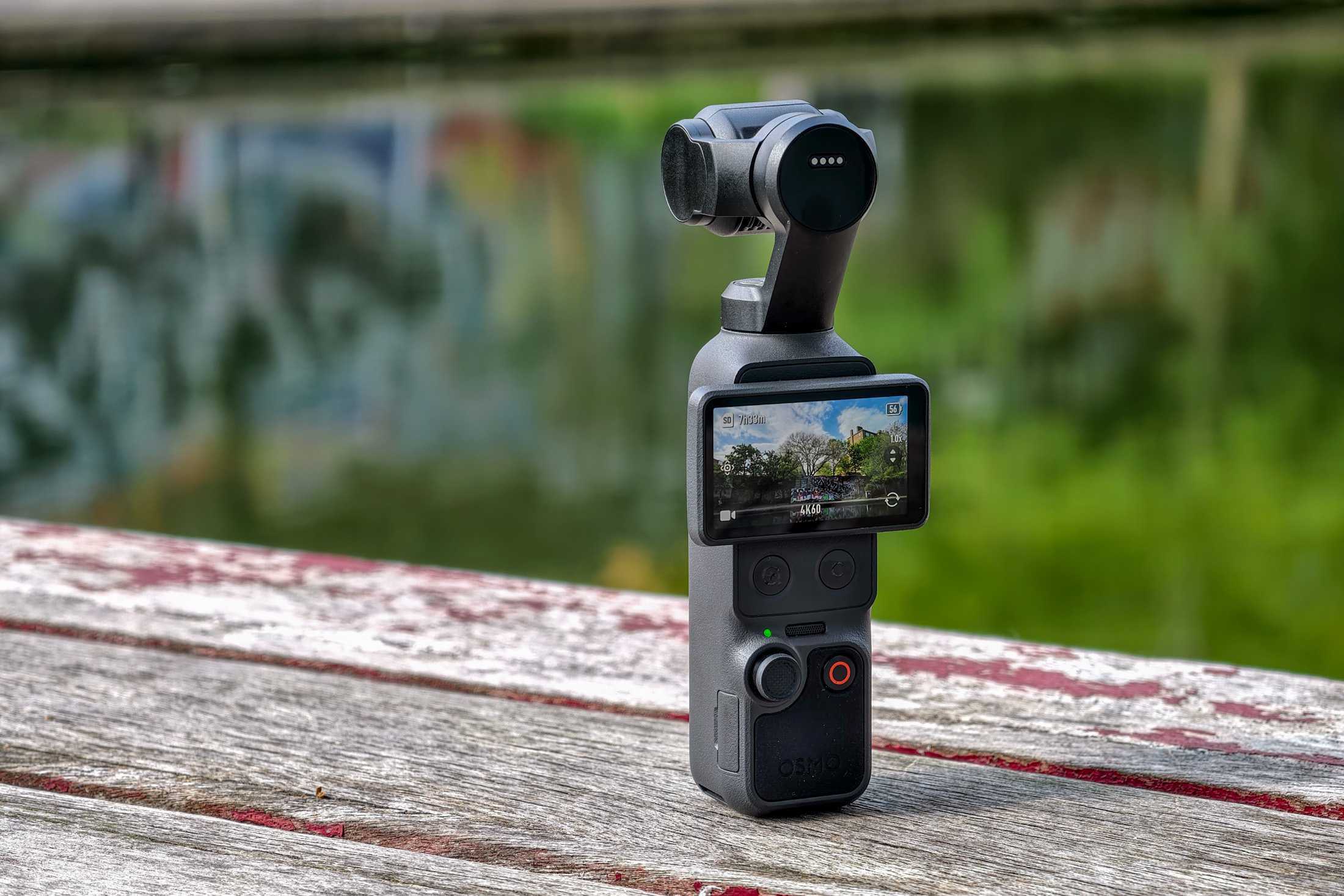 Photo of the DJI Osmo Pocket 4 standing on a wooden table in front of a canal, showing the screen with the camera view