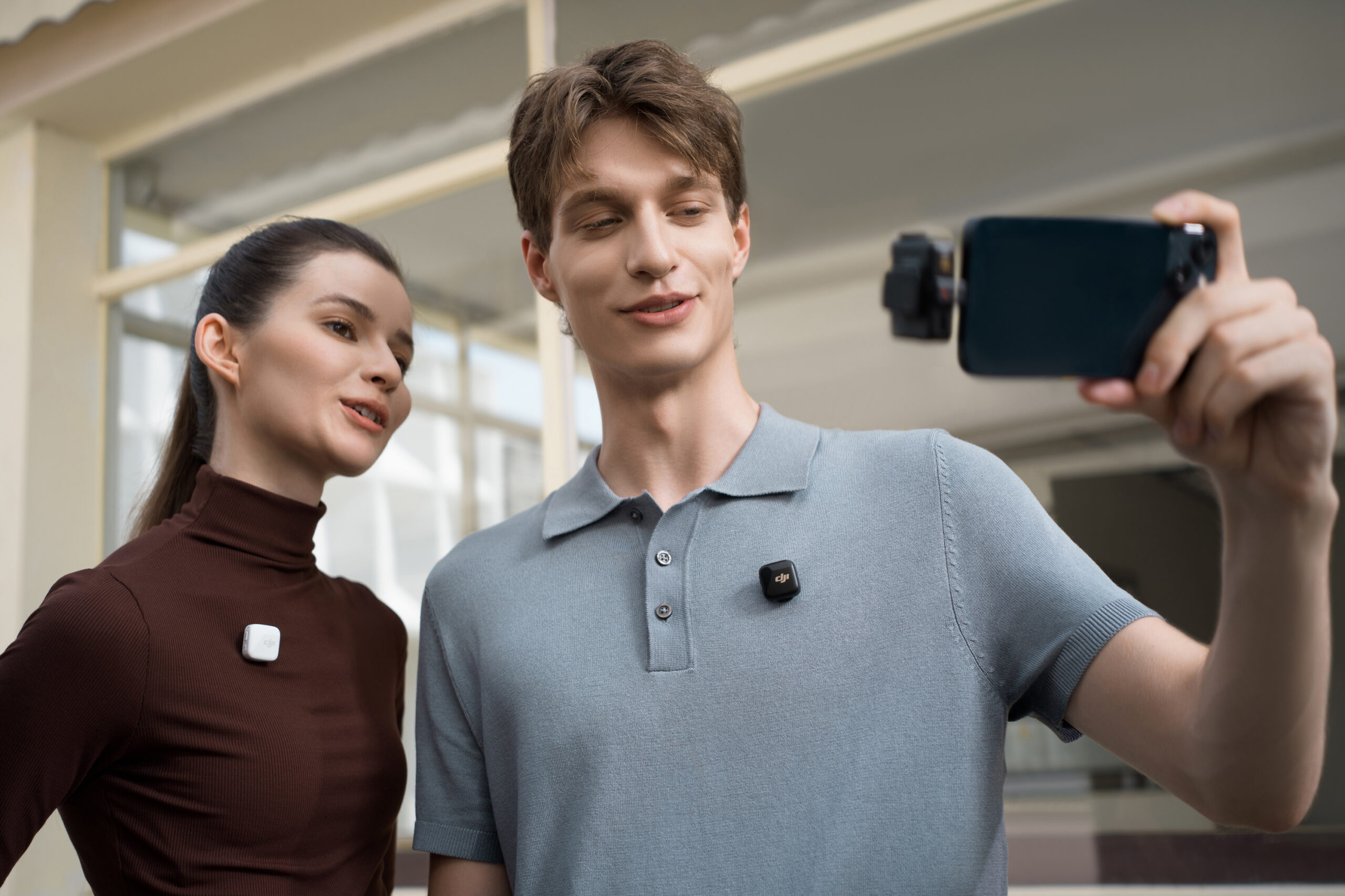 Two people wearing the DJI Mic Mini transmitters attached to their shirts while one holds a smartphone with a wireless receiver attached.