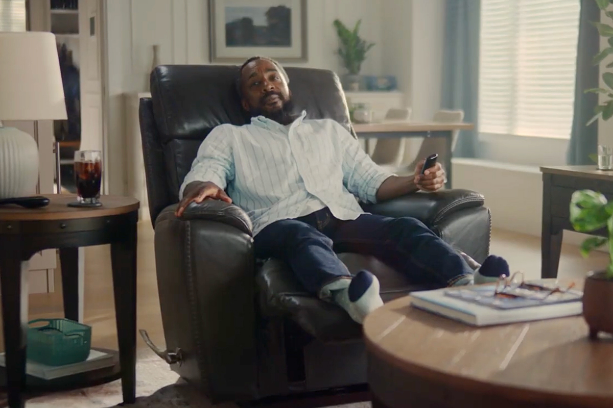A person sits in a La-Z-Boy recliner in a living room with a TV remote in hand.