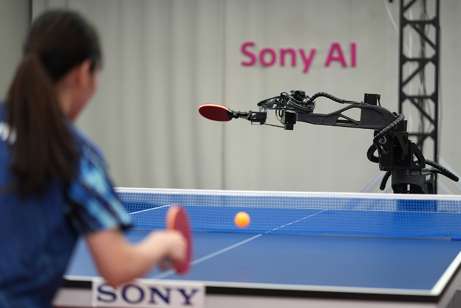 A human table tennis player out of focus in the foreground competing against a Sony’s Ace robot holding a red paddle.