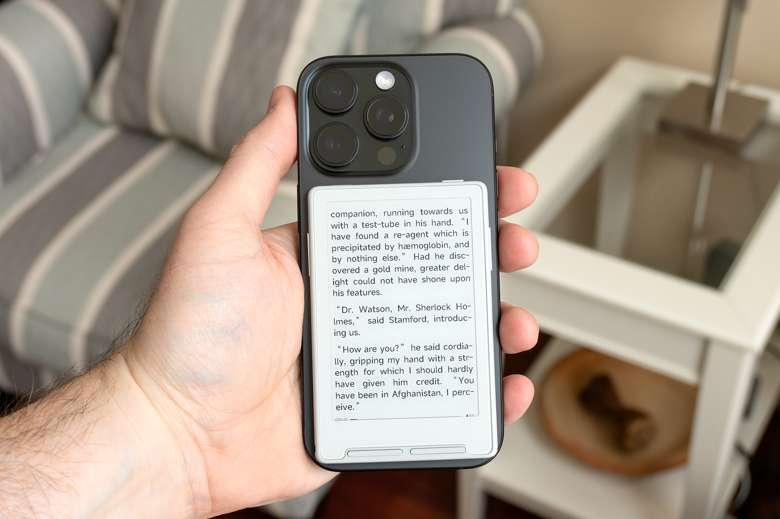 A person holds an iPhone with the Xteink X3 e-reader attached to the back showing an ebook on screen.