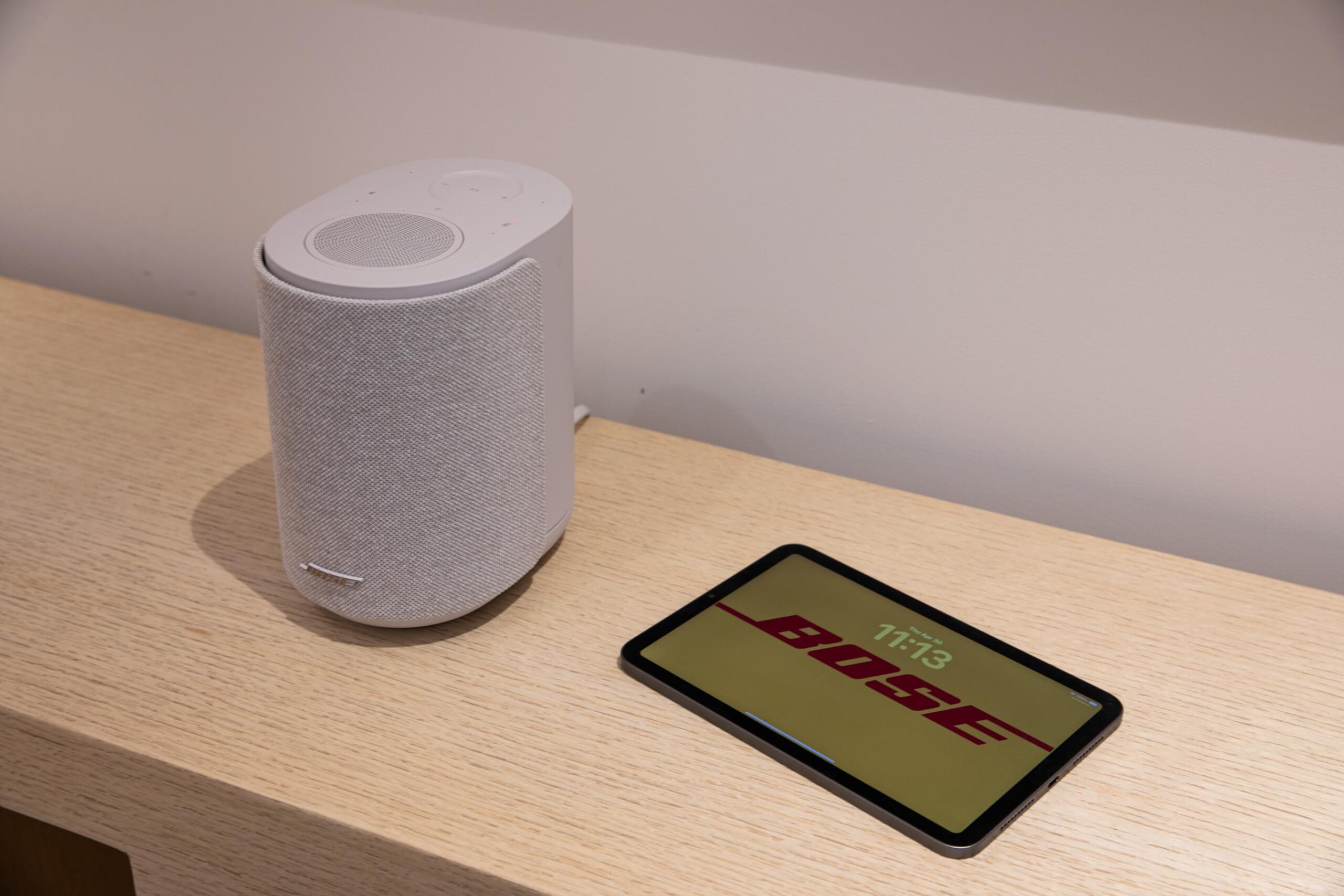 The Bose Lifestyle Ultra Speaker on a wooden table with a tablet displaying the Bose name next to it.