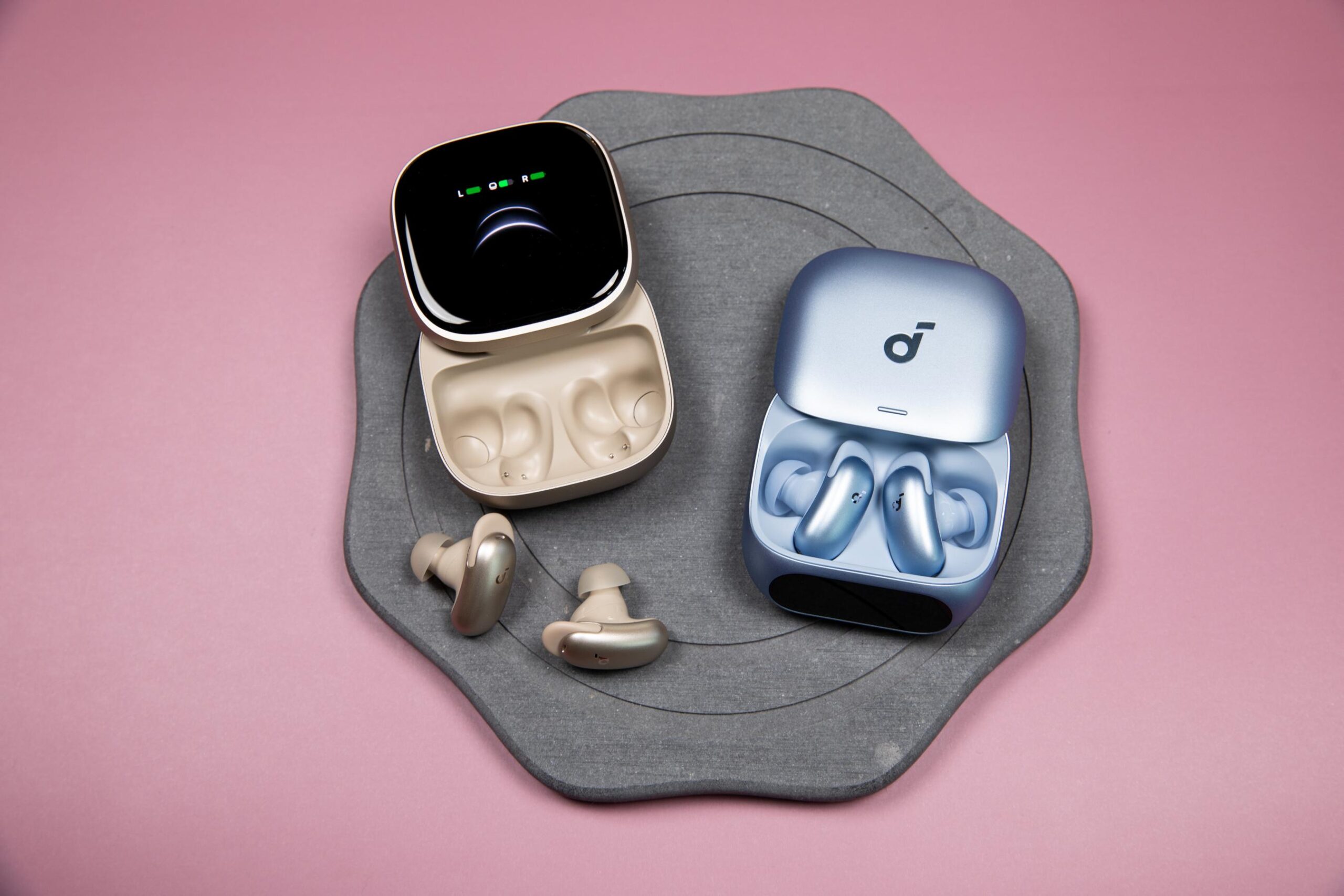 Champagne Soundcore Liberty 5 Pro Max earbuds and their case next to blue Soundcore Liberty 5 Pro earbuds on a stone trivet with a mauve-colored background.