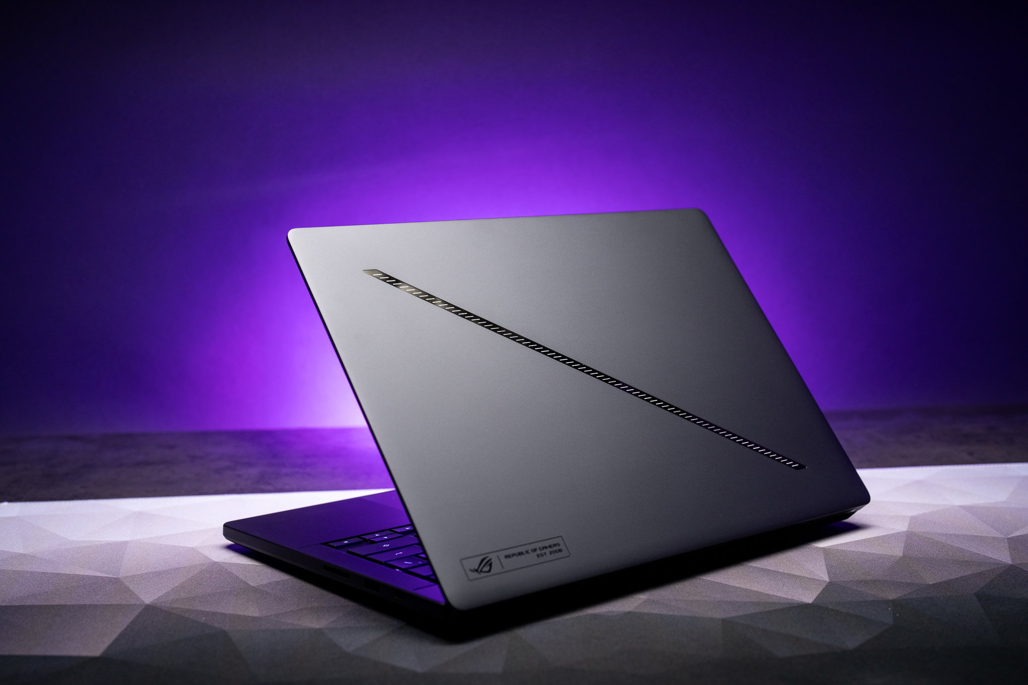 A rear view of an Asus ROG Zephyrus G14 (2026) laptop on a desktop with purple light behind it in the background.