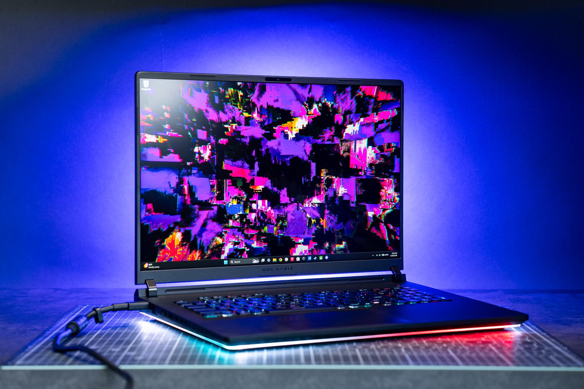 A pre-production 2026 Asus ROG Strix Scar 18 gaming laptop sitting on a black cut mat in front of a blue illuminated backdrop. The large black laptop is emanating colorful lights from its keyboard and underglow around its chassis.
