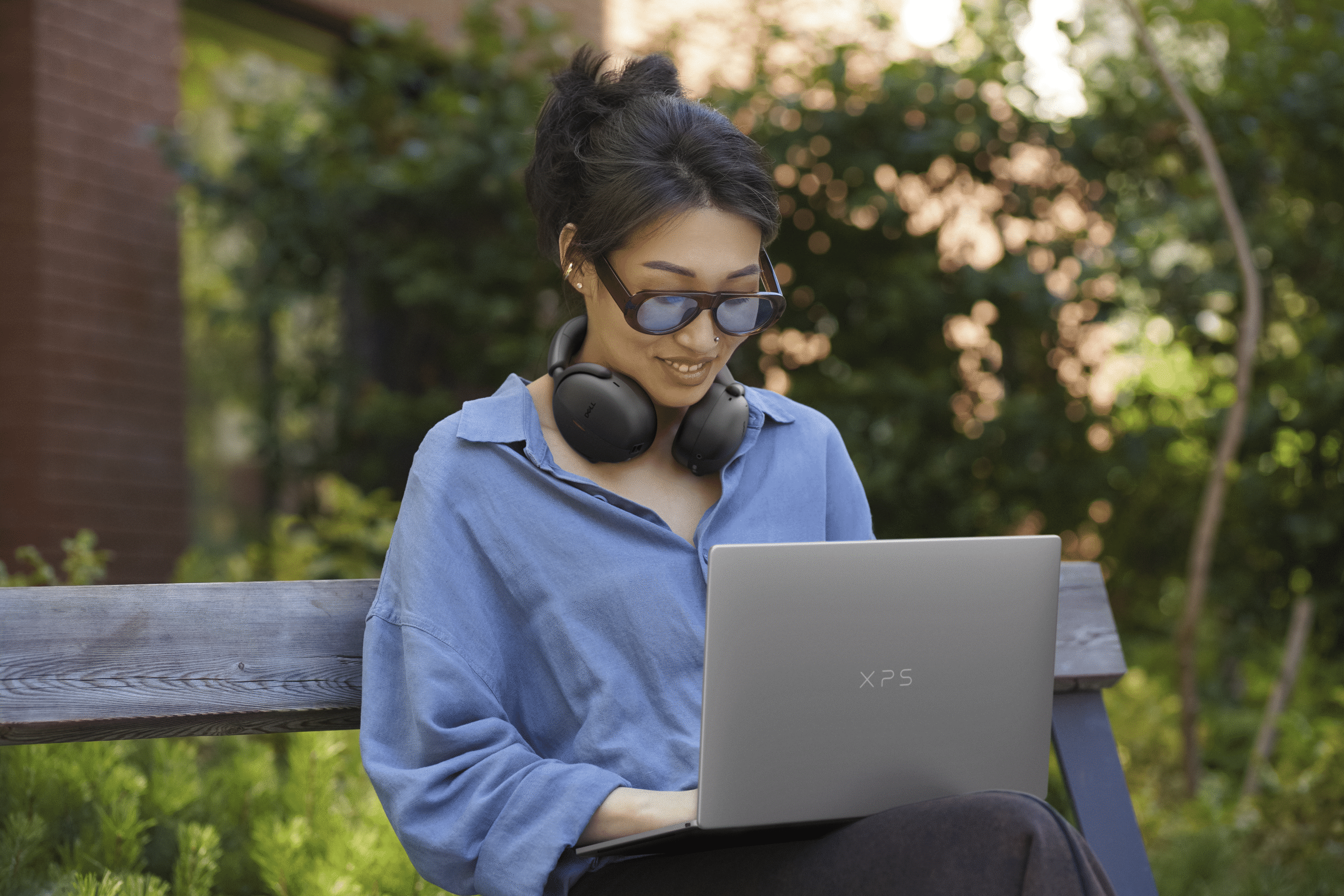 A student sitting on a park bench while wearing headphones around their neck, working on a Dell XPS 13 laptop.