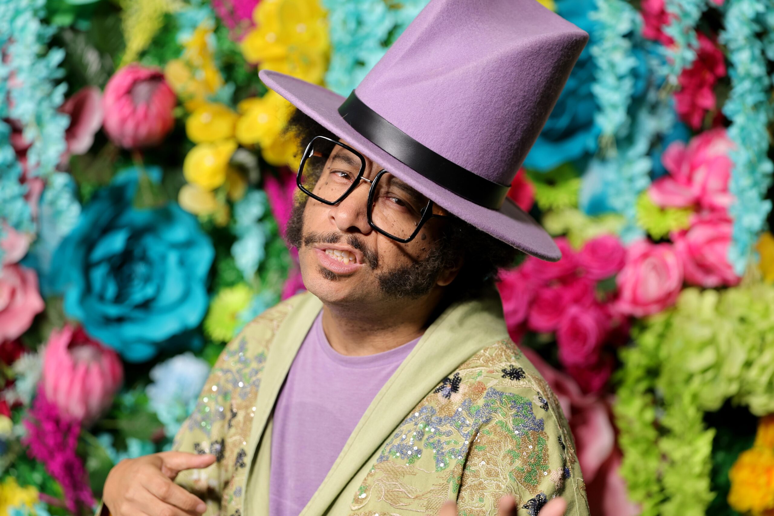 A man wearing a purple shirt, a sage cardigan, glasses, and a large purple hat. Behind the man is a wall of pastel flowers.
