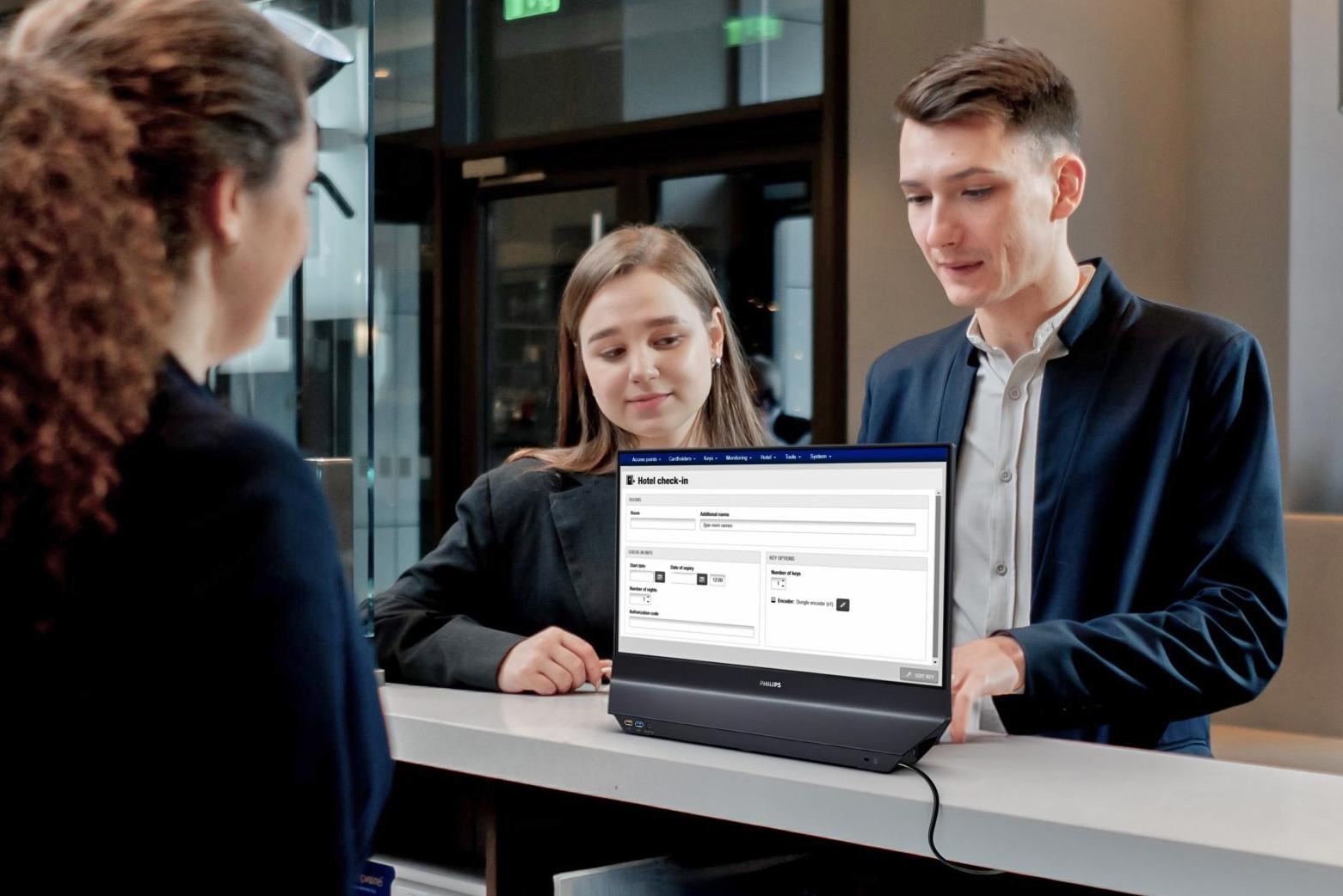 Two people at a front desk look at one side of a monitor while an employee looks at the other.