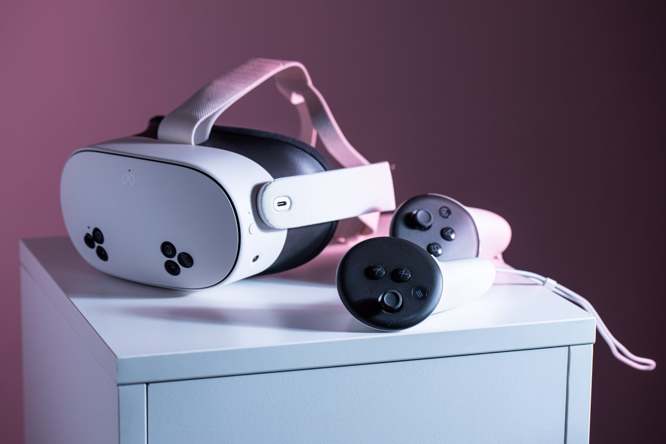 A white VR headset with a three-lens cluster on the lower left and right of the front visor, looking like a sad robot, plus a pair of handheld controllers with white grips and black control surfaces, on a small white end table, with pinkish background and lighting.