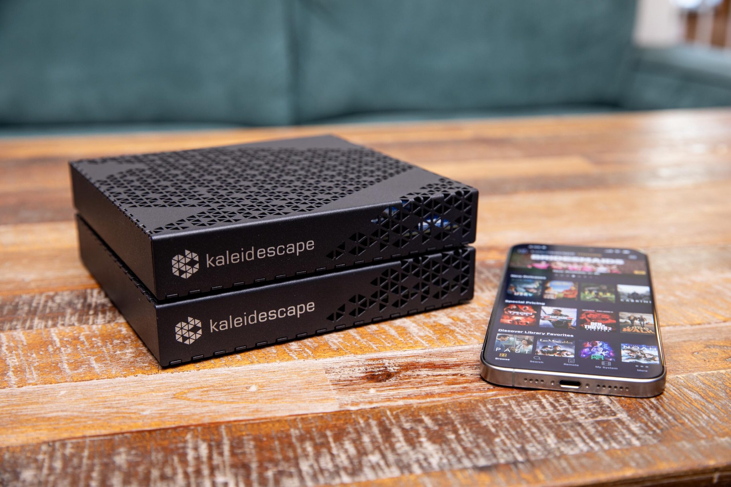 A Kaleidescape Strato E player a Mini Terra Prime server stacked next to an iPhone displaying the Kaleidescape app on a wooden table.