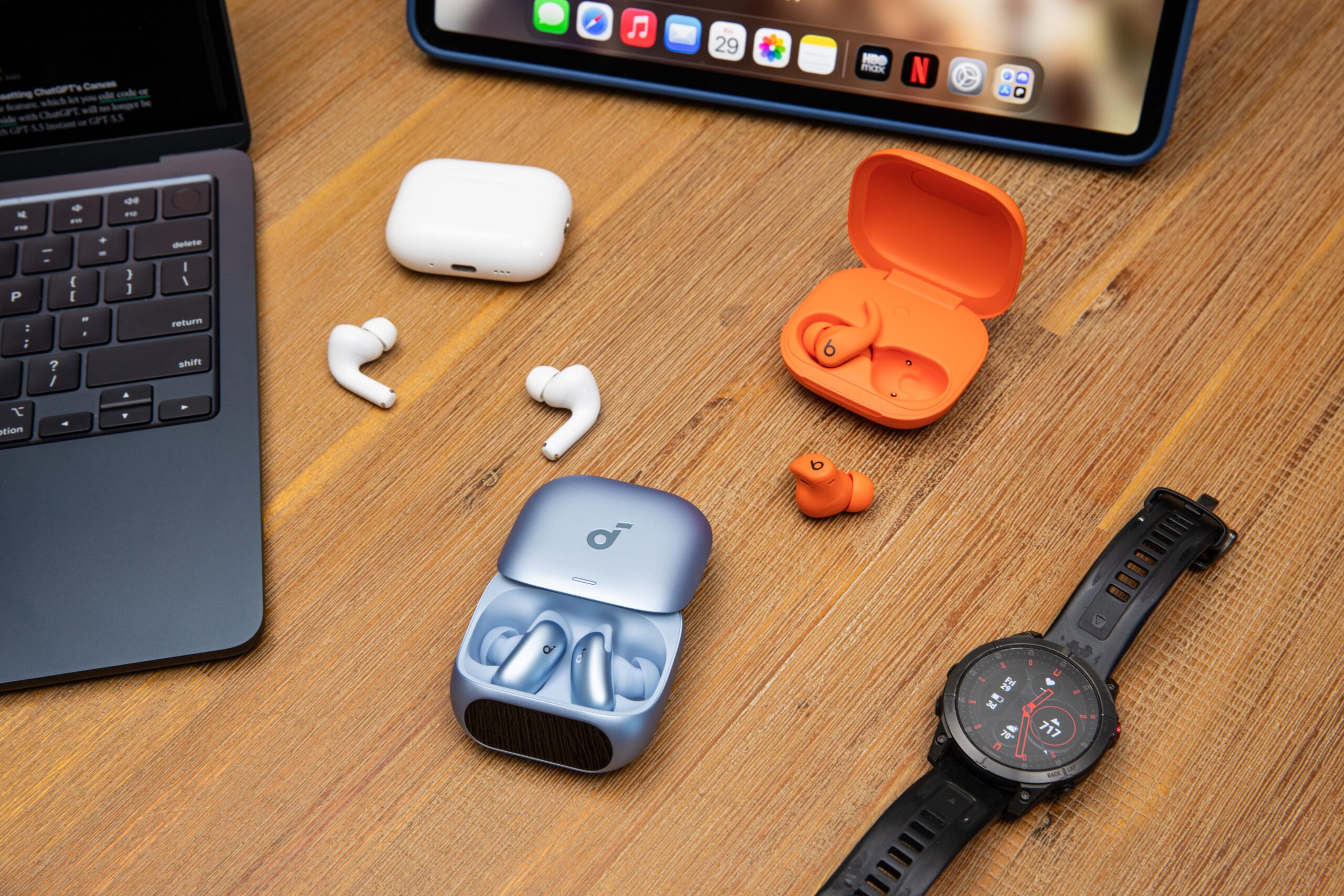 The Apple AirPods Pro 3, Beats Powerbeats Fit, and Soundcore Liberty 5 Pro on a wooden table between a MacBook, iPad, and Garmin Epix watch.