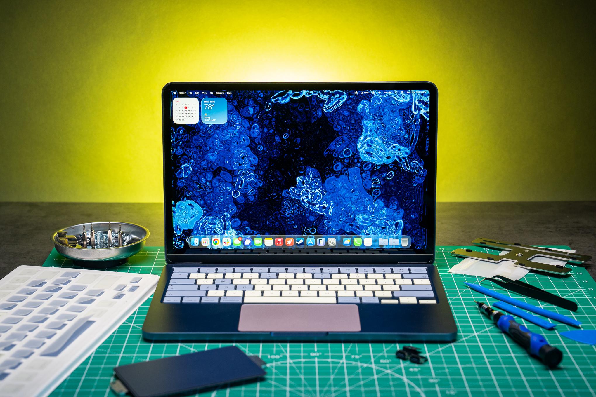 An indigo MacBook Neo with color-mixed parts sits on a green cut mat besides tools and spare parts. The laptop’s trackpad is blush, and its keys are a mix of citrus and the original indigo.