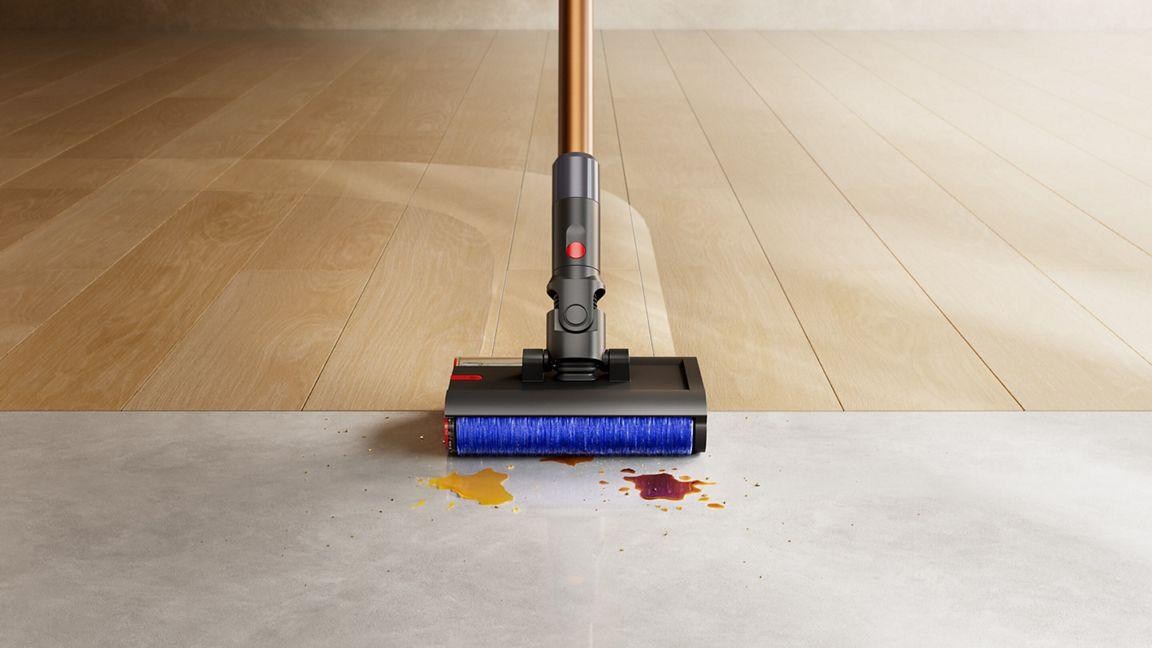 A Dyson PencilWash cordless hard floor cleaner cleaning a path across a wood floor and about to go over two small spills on an adjacent section of tile.