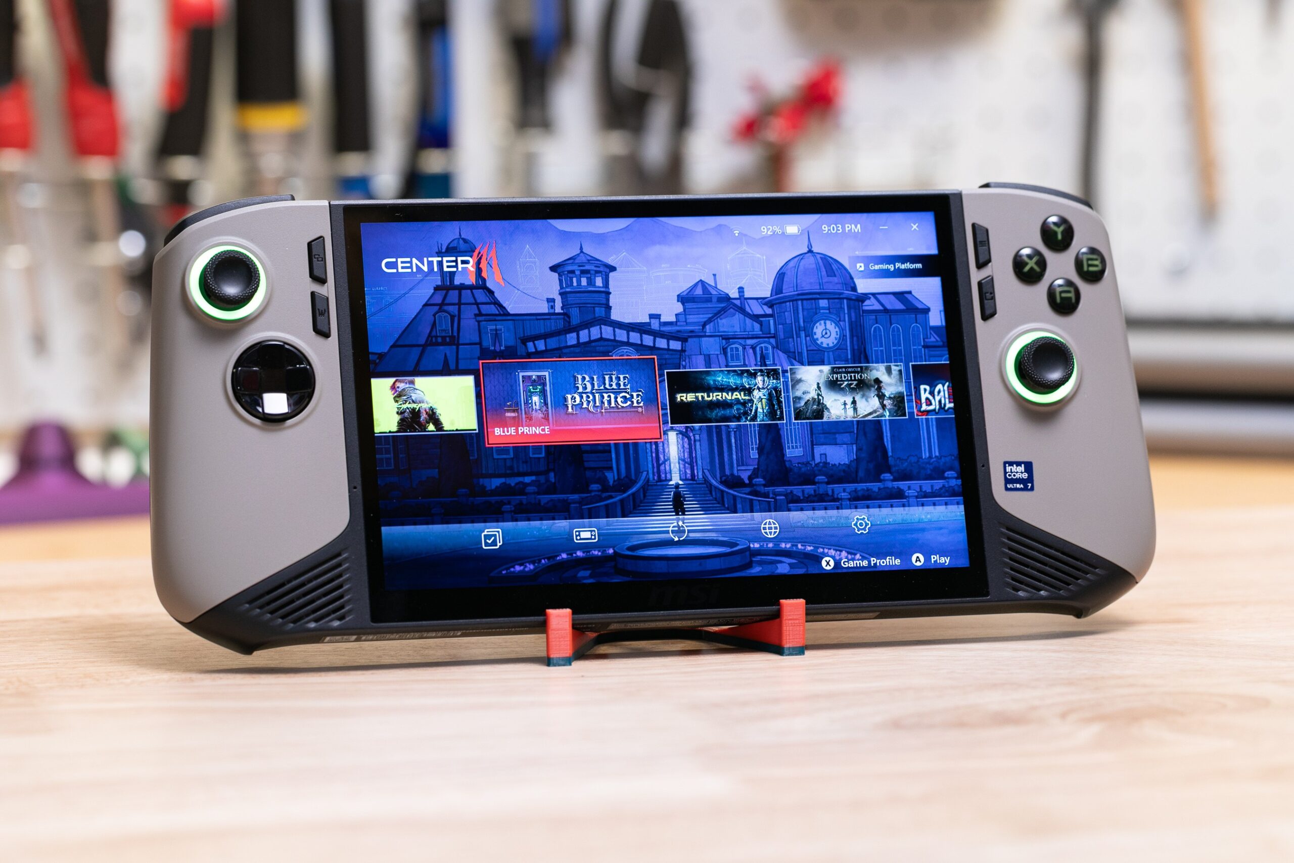 A gray handheld gaming PC, with black accents and RGB lit joysticks and face buttons, with a screen showing Blue Prince on it.
