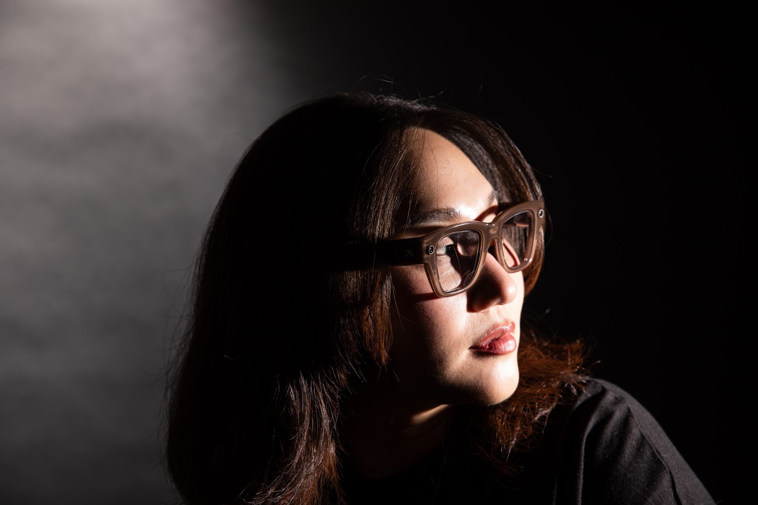 Senior reviewer Victoria Song wearing Meta Ray-Ban Display glasses while looking over her shoulder. The photo is lit so there are dramatic shadows across her face.