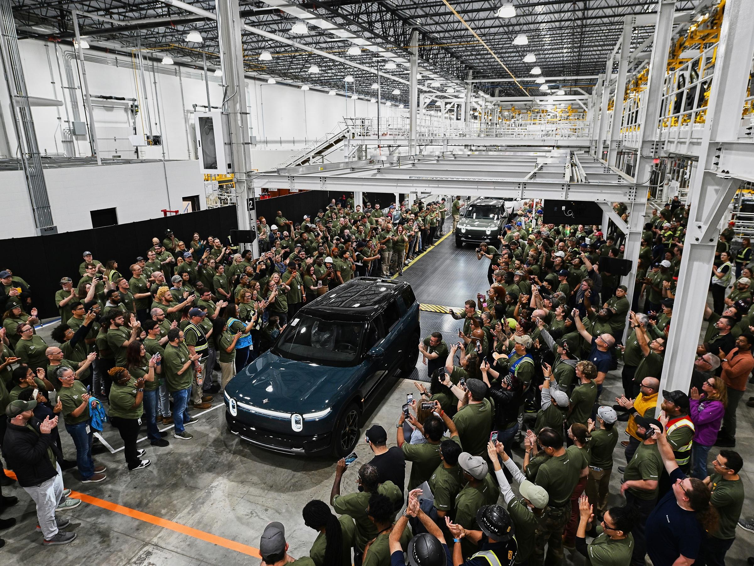 Wide view of a dark blue Rivian driving down a narrow path of the factory floor with people gathered on either side.