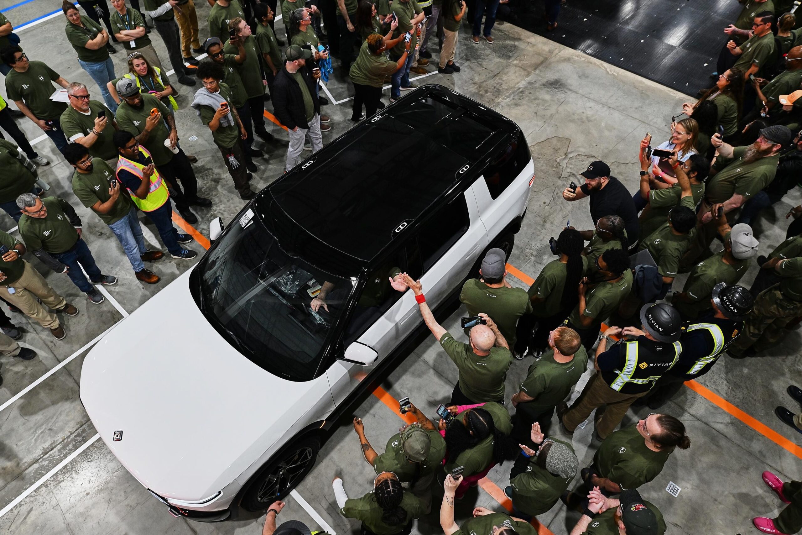 People lined up on either side of a white Rivian driving down a narrow path in a factory. The driver gives a high five to a person taking pictures outside of the vehicle. People lined up on either side of a white Rivian driving down a narrow path in a factory. The driver gives a high five to a person taking pictures outside of the vehicle.