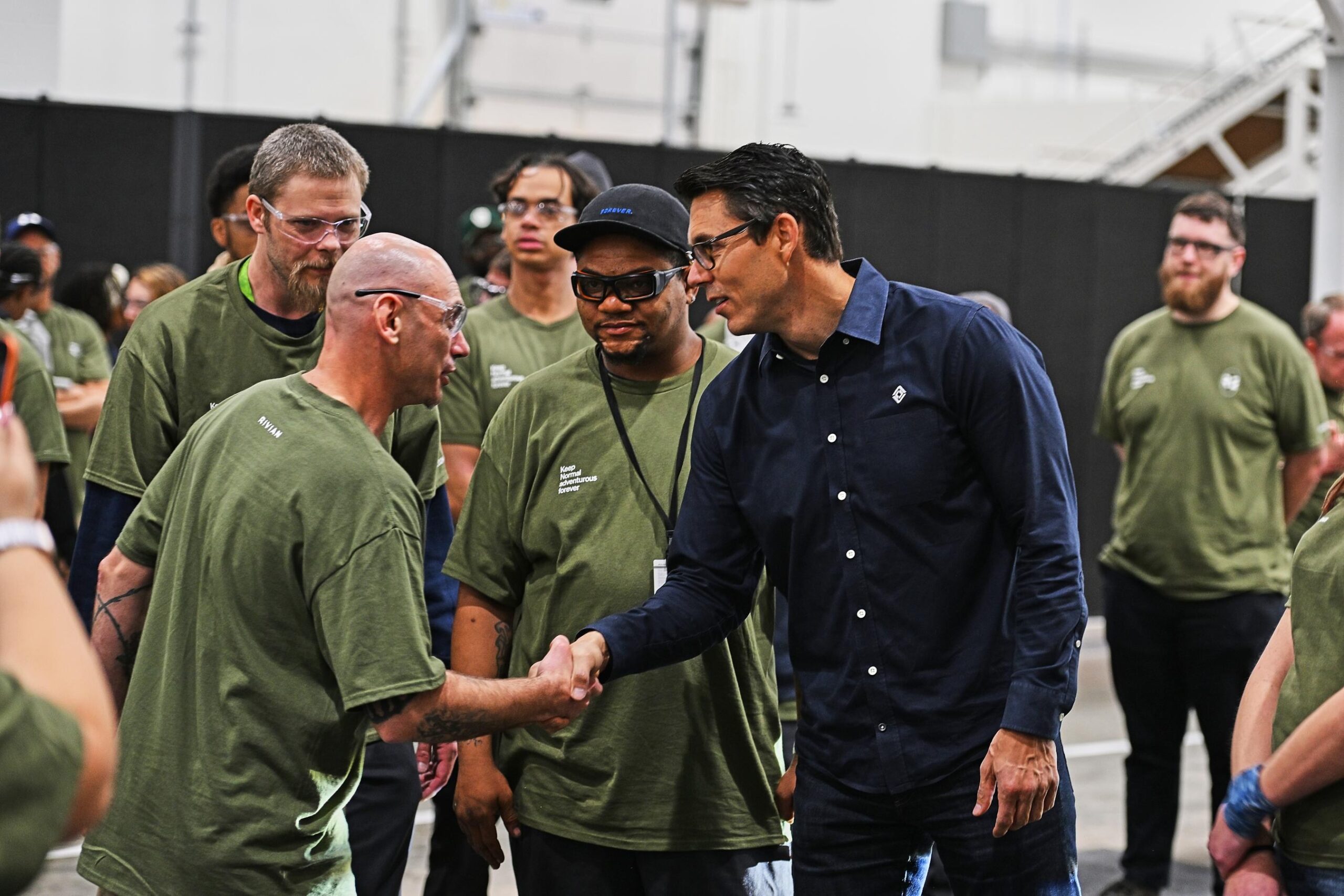 CEO RJ Scaringe in a blue long sleeved shirt and glasses is shaking the hand of a man in safety goggles and a green t-shirt. A group of people also in green t-shirts are around them. CEO RJ Scaringe in a blue long sleeved shirt and glasses is shaking the hand of a man in safety goggles and a green t-shirt. A group of people also in green t-shirts are around them.