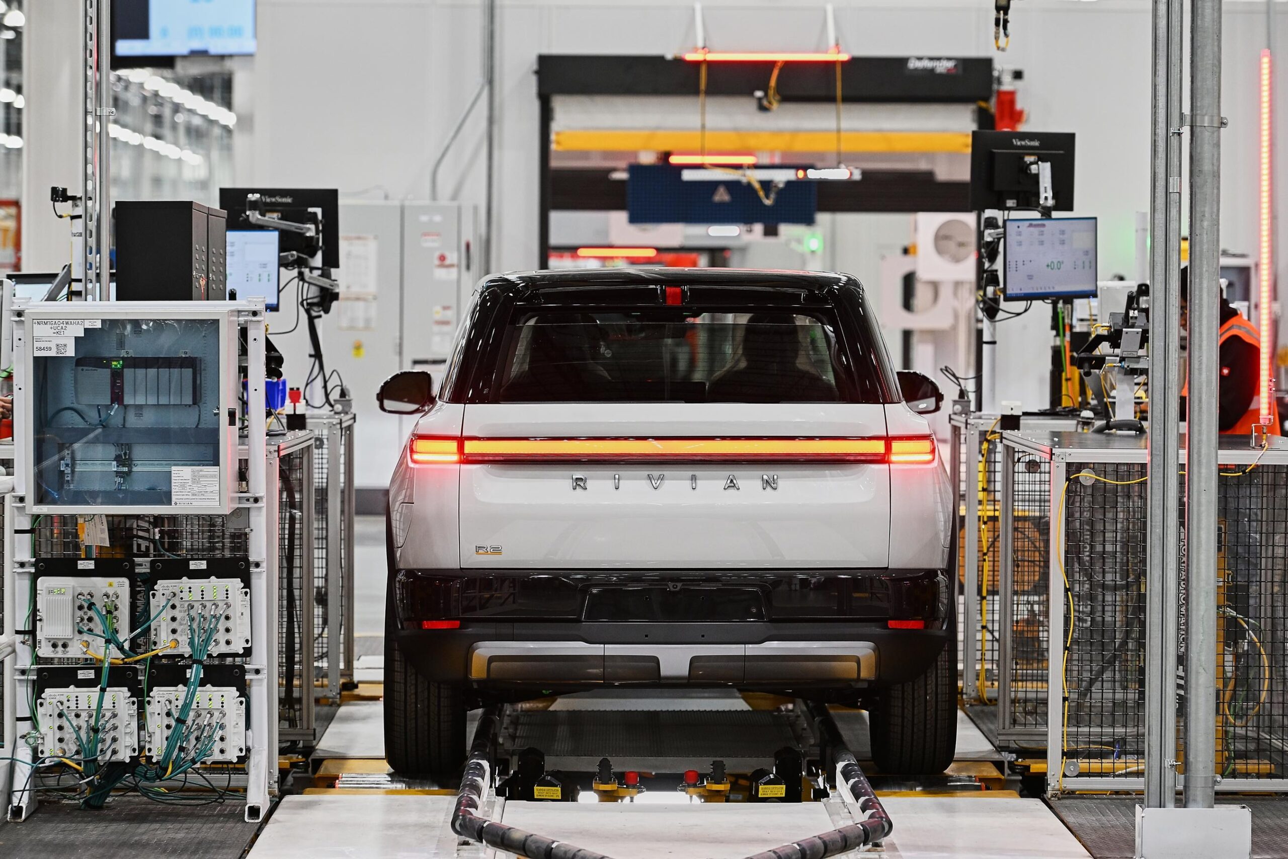 A white Rivian in between computer equipment seen from behind at the factory. A white Rivian in between computer equipment seen from behind at the factory.
