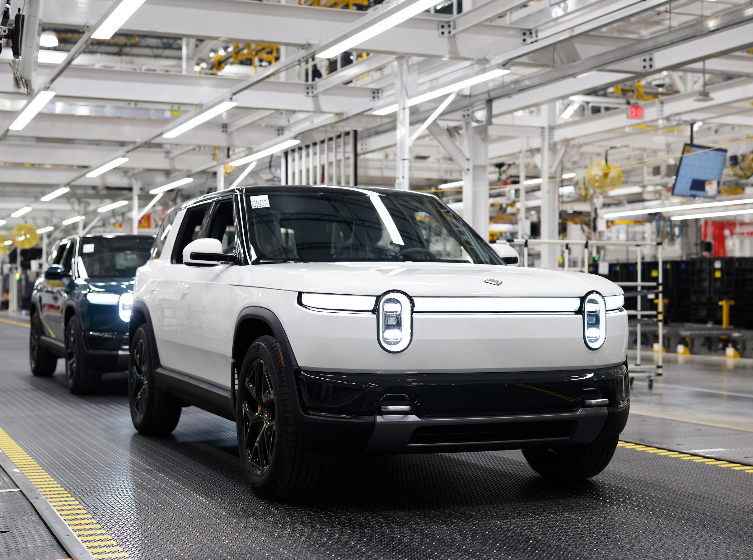 Two Rivian R2's with headlights on are displayed, the first is white and the one behind is blue. Two Rivian R2's with headlights on are displayed, the first is white and the one behind is blue.