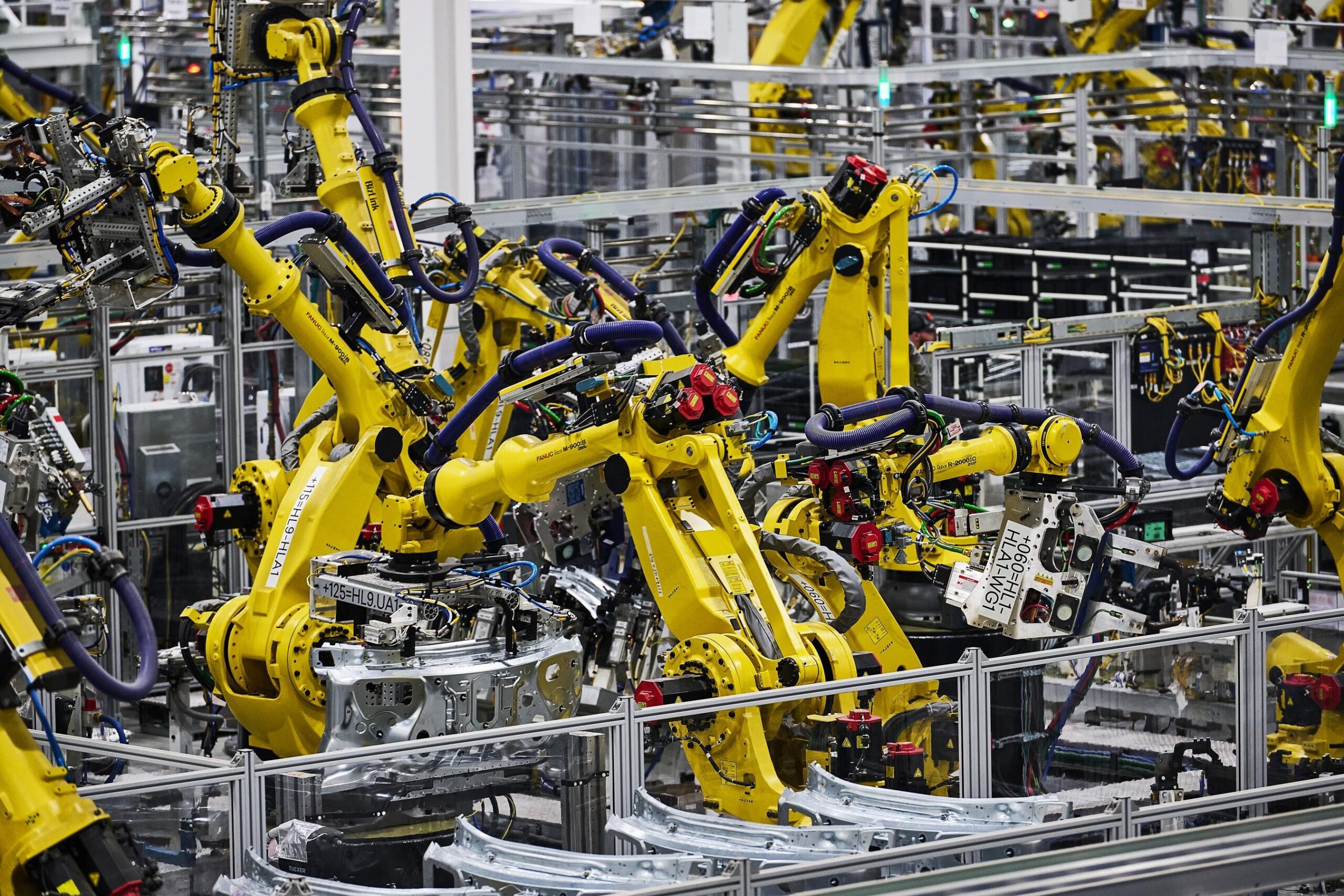 A cluster of yellow machine arms working on unspecified silver car parts on the Rivian factory floor. A cluster of yellow machine arms working on unspecified silver car parts on the Rivian factory floor.