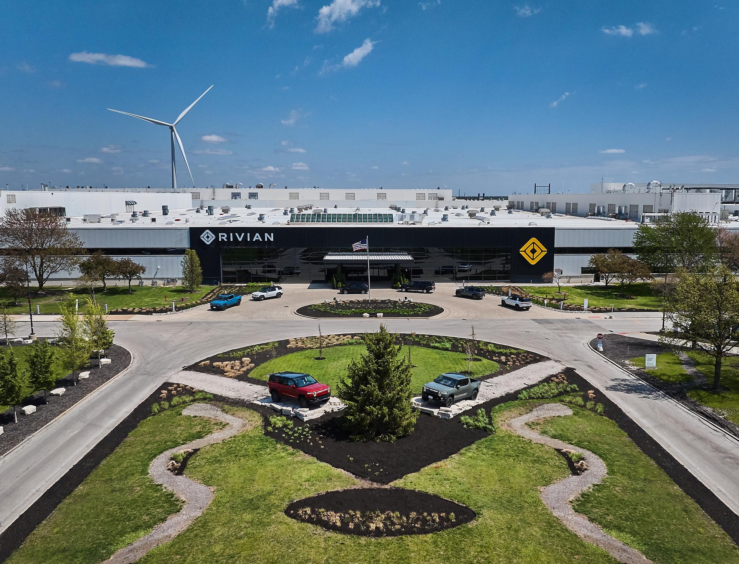 A drone shot of the entrance of the Rivian factory. There's a manicured grassy area with two vehicles displayed along the road leading to the entrance of the enormous Rivian building. It's a bright sunny day with a blue sky overhead. A drone shot of the entrance of the Rivian factory. There's a manicured grassy area with two vehicles displayed along the road leading to the entrance of the enormous Rivian building. It's a bright sunny day with a blue sky overhead.