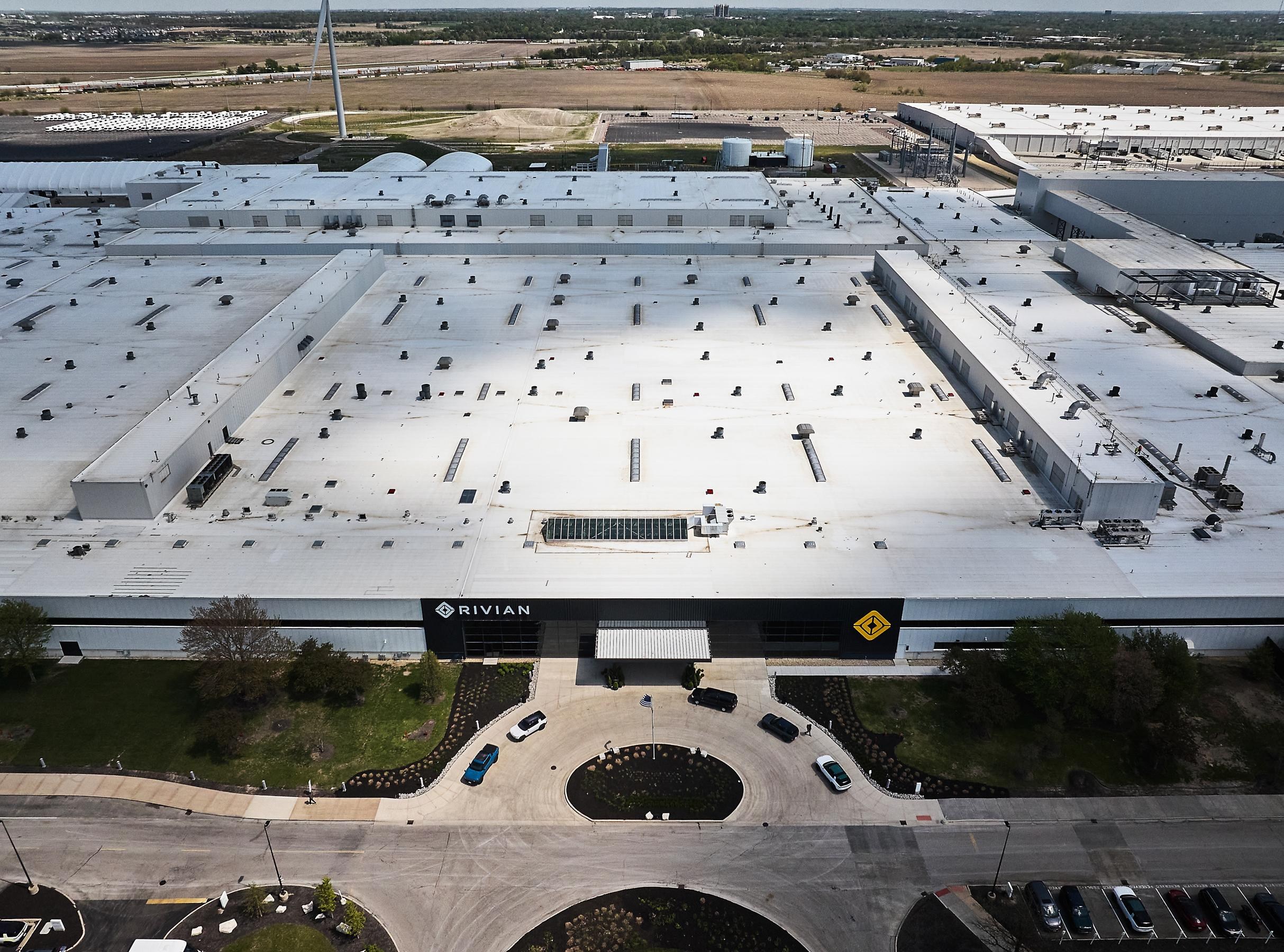 An aerial view of Rivian's huge factory with rural land seen behind it and cars parked in its circular driveway in front. An aerial view of Rivian's huge factory with rural land seen behind it and cars parked in its circular driveway in front.
