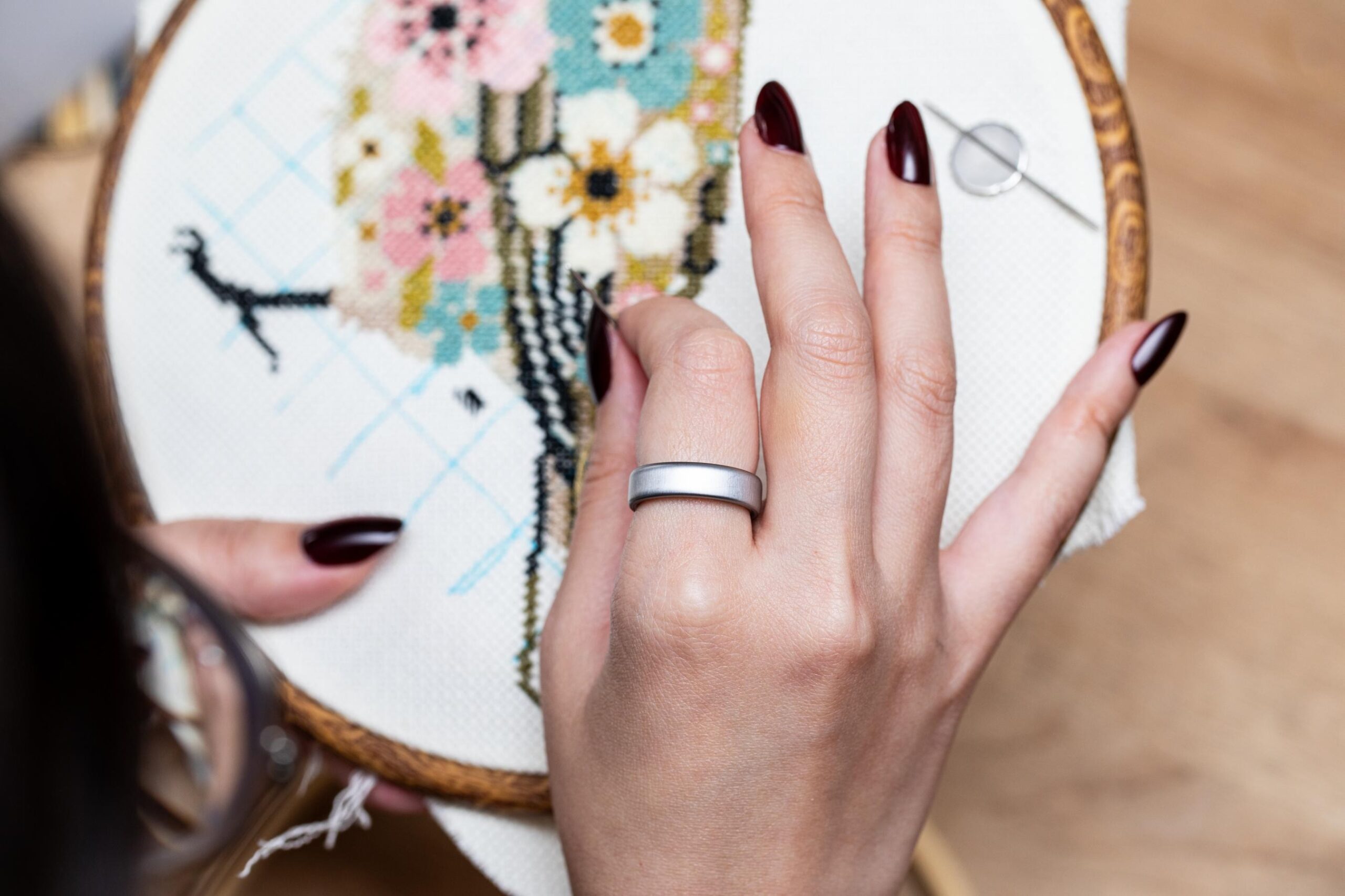 Close up of an Oura Ring 5 on someone’s finger as they work on a cross stitch project of a bird with florals. 