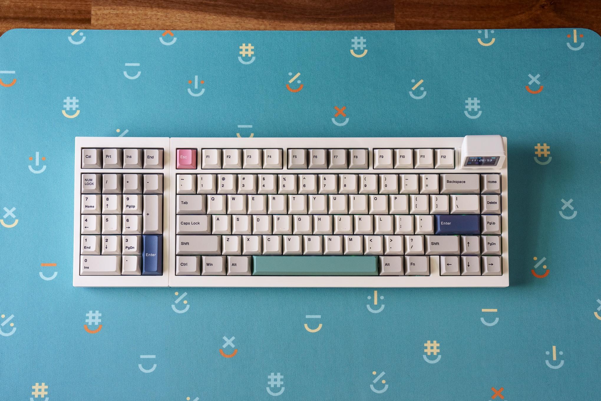 A top-down view of an Epomaker RT98 mechanical keyboard on a colorful desk mat with a repeating emoji pattern. The beige keyboard with multicolored keycaps has an 1800 layout with a southpaw-oriented number pad.