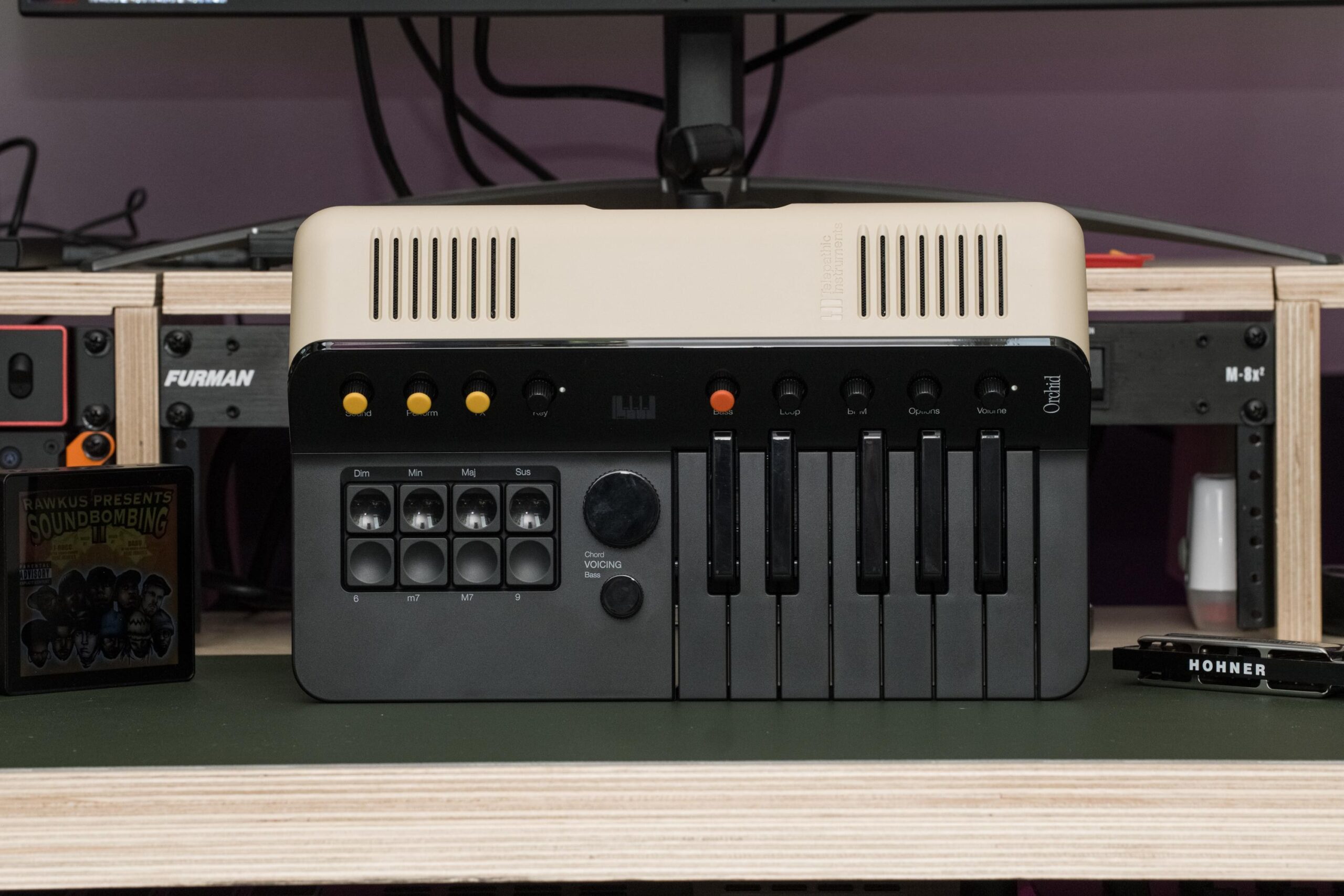 The Telepathic Instruments Orchid in all its tan and black glory on a desk next to the Sleevenote media player, with Rawkus Records Soundbombing II queued up on it, and a harmonica on the other side, because the Orchid is a tribute to electric chord organs of the past, which are basically just giant harmonicas hooked up to a fan.