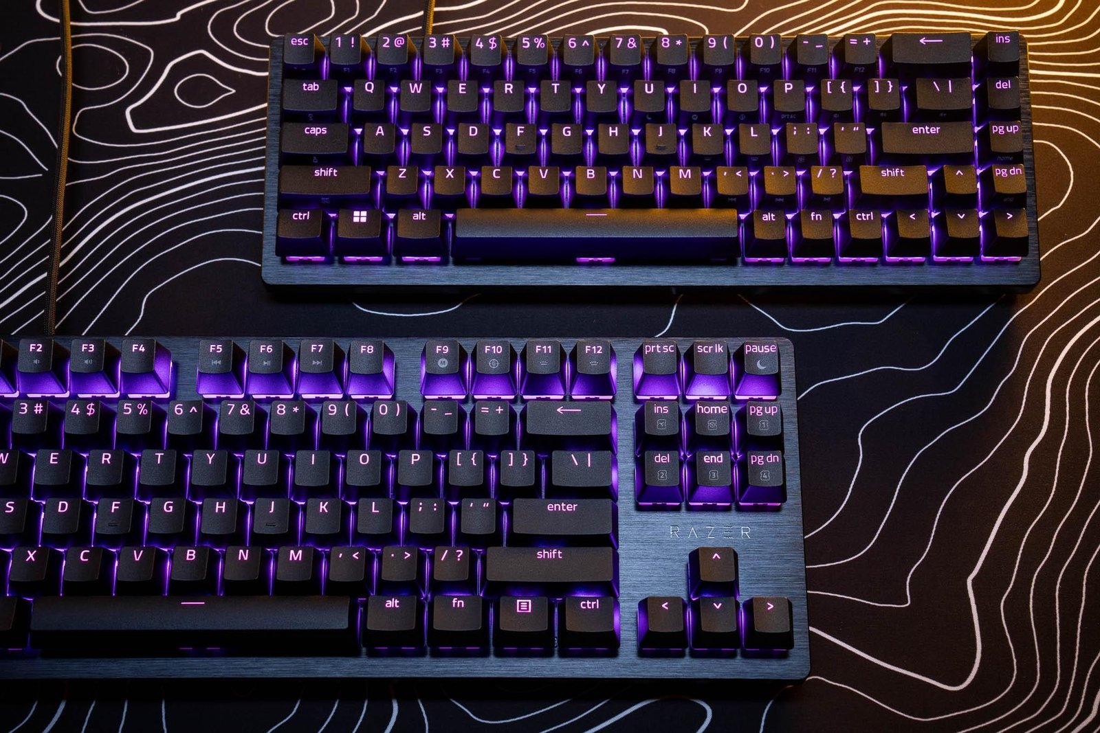 A Razer Huntsman V3 HE Magnetic TKL keyboard (lower-left) and a Huntsman V3 HE Magnetic Mini 65-percent keyboard (upper-right) on a desk mat.