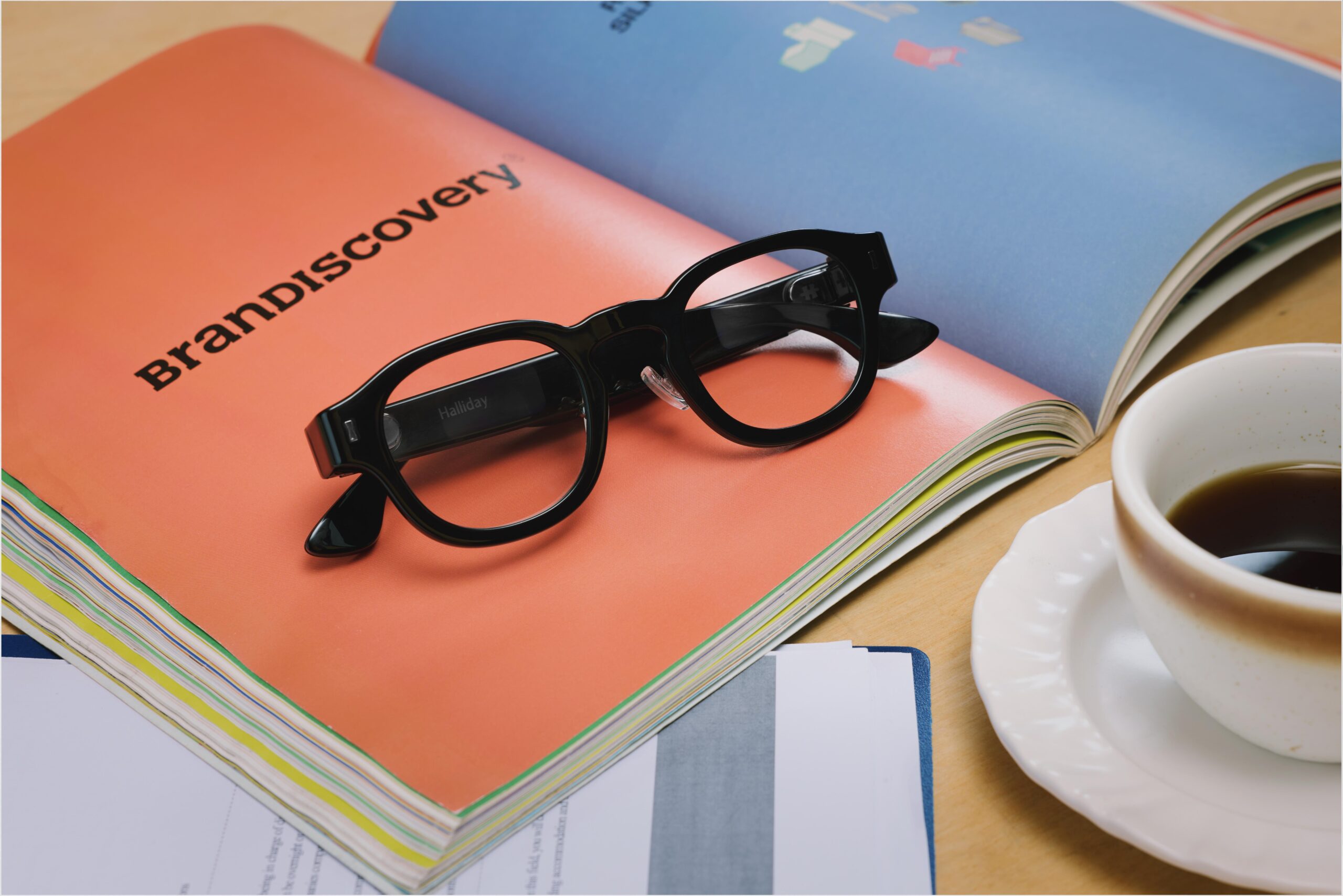 The Halliday Gen 2 glasses sit on top of an open magazine with a cup of coffee visible in the corner.