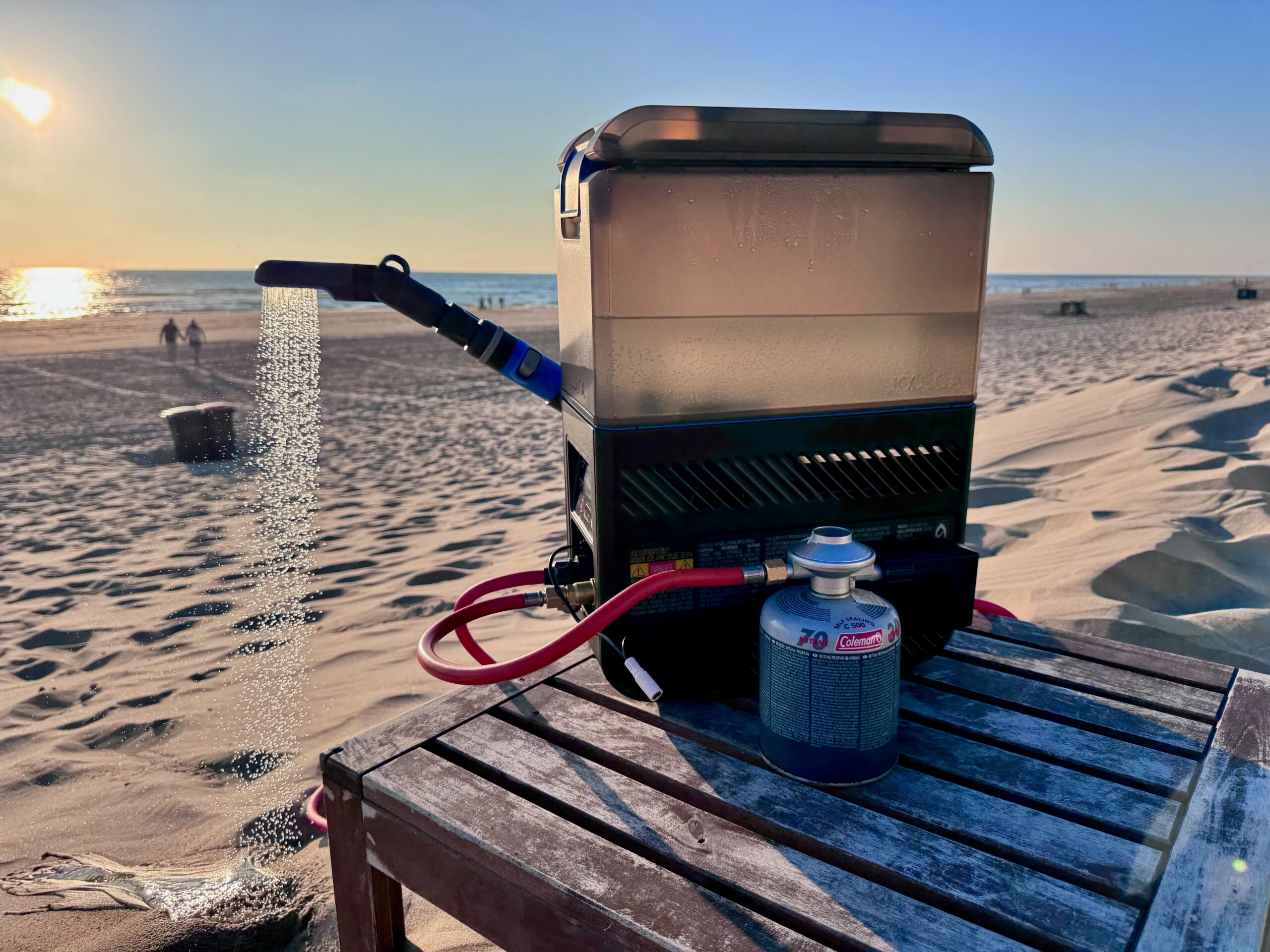A beach scene showing the Joolca Hottap Go on a low table sitting in the sand at sunset with the water flowing out of the showerhead magnetically attached to the side of the jerrycan-like chassis.