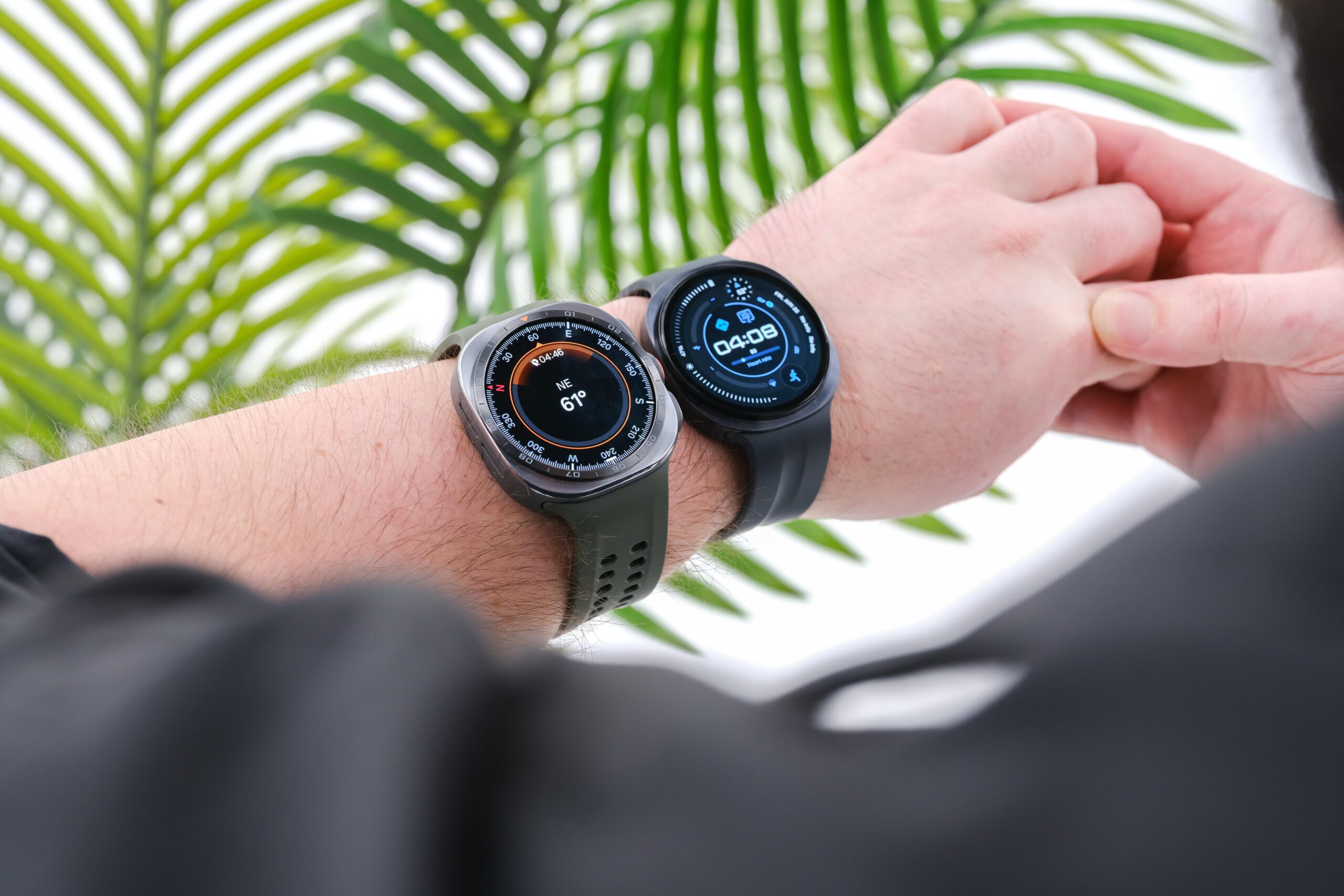 An over-the-shoulder view of the Samsung Galaxy Watch Ultra 2 and Galaxy Watch 9 on a man’s wrist with green plant fronds in the background.