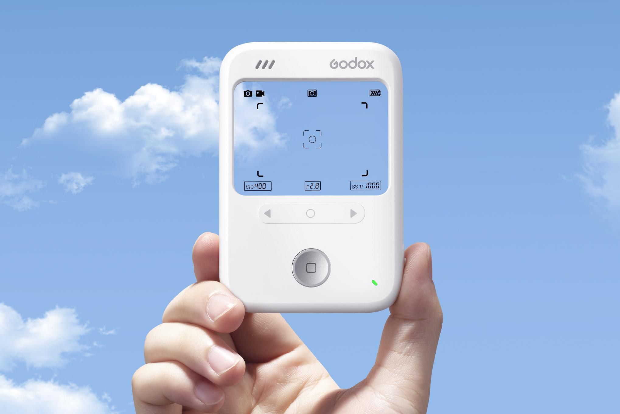 A hand holds the Godox C100 digital camera with a blue cloudy sky in the background.