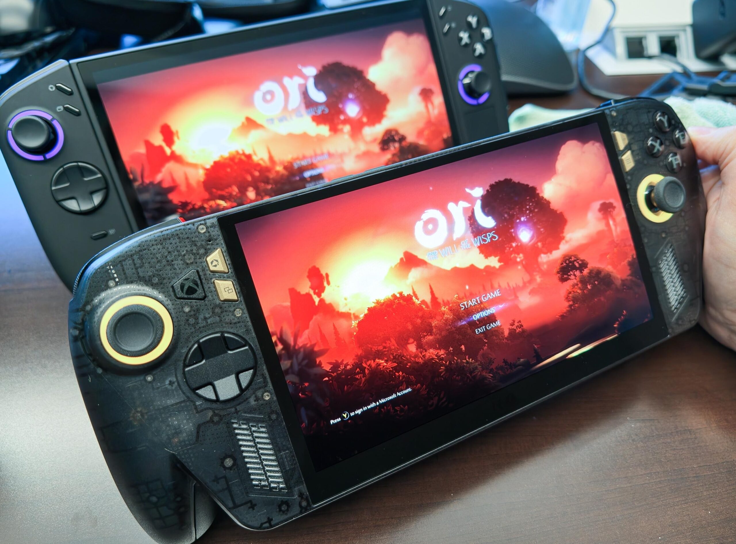Two OLED screen handhelds face the camera, with Ori and the Will of the Wisps’ golden sunset on their screens. The front handheld, the Ally X20, has a translucent shell.