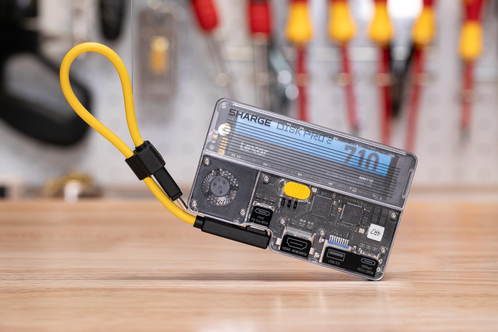 A playing-card-deck-sized gadget with a transparent front, showing off computer chips and a fan and an SSD inside, as well as a detachable yellow USB-C cable shaped into a lanyard.
