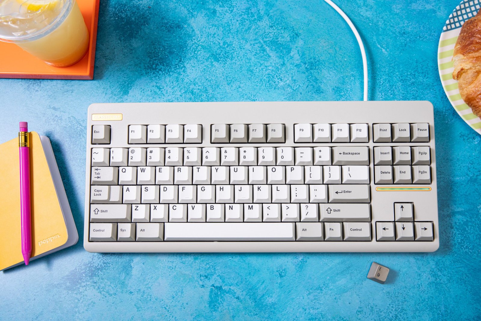 A beige keyboard with cream-and-beige keycaps on a blue watercolor deskmat, surrounded by objects including a yellow notepad and pink pen.
