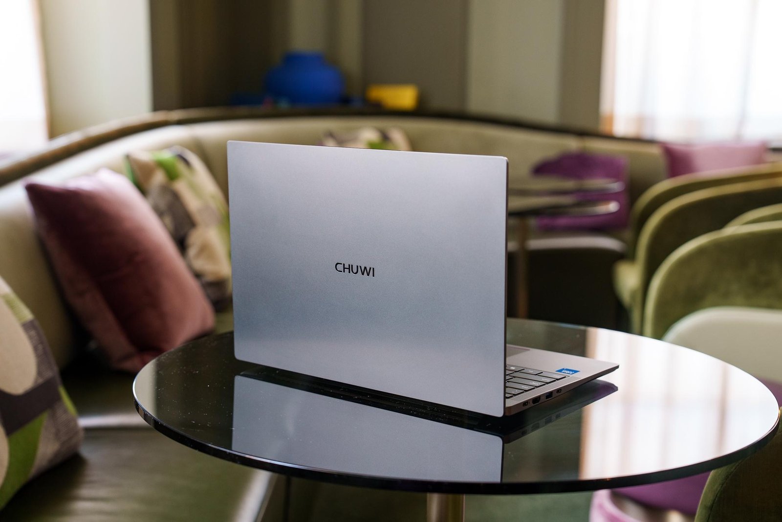 A Chuwi UniBook laptop, facing away from the camera to show its lid. It’s sitting on a glss table admist a plush meeting space filled with colorful couches and chairs.
