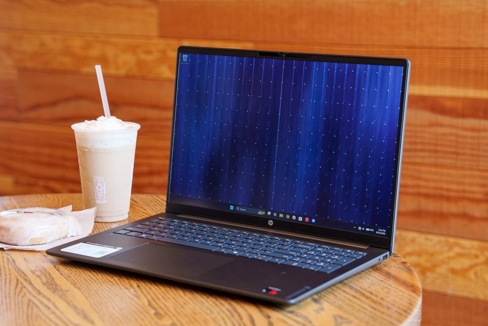 An HP OmniBook 3 16 laptop on a wood table next to a frozen coffee drink and pastry.