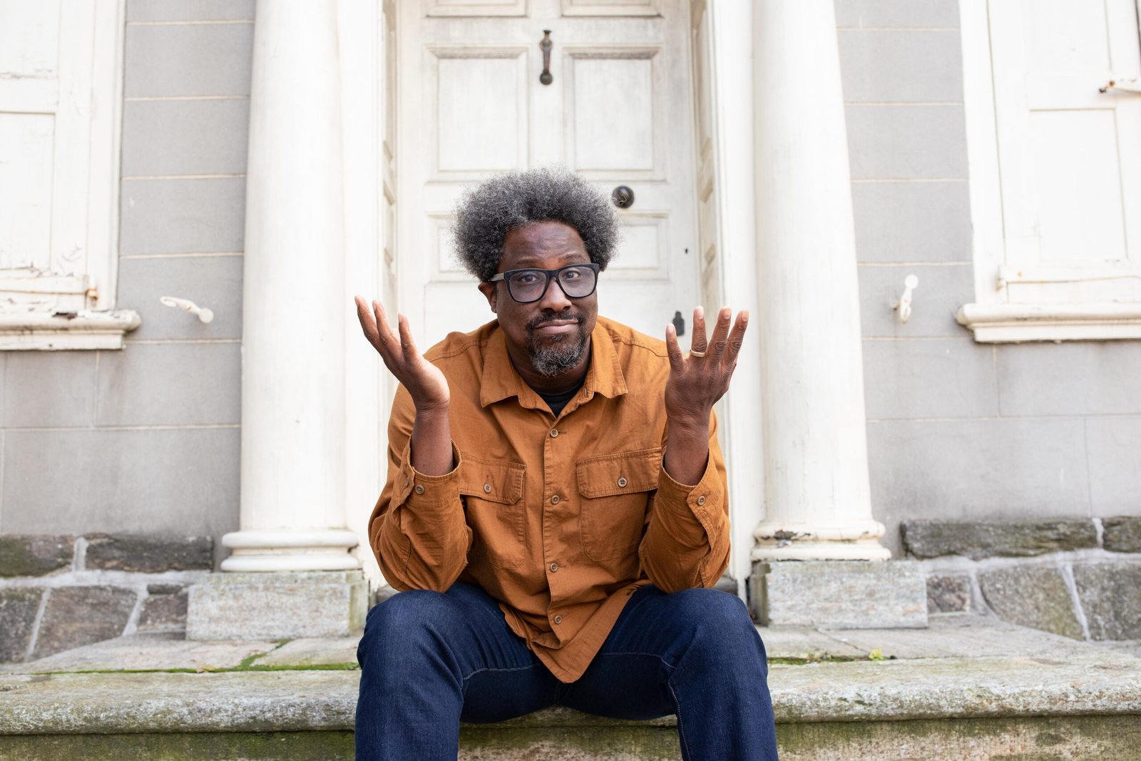 W. Kamau Bell looking confused sitting on the steps outside of a white door.