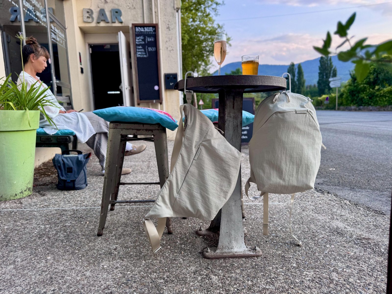 Peak Design’s new crossbody bag and backpack hanging off a round table outside a bar. There are mountains in the distance and a glass of beer and wine sit on the table. A woman sits on a bench off to the side.