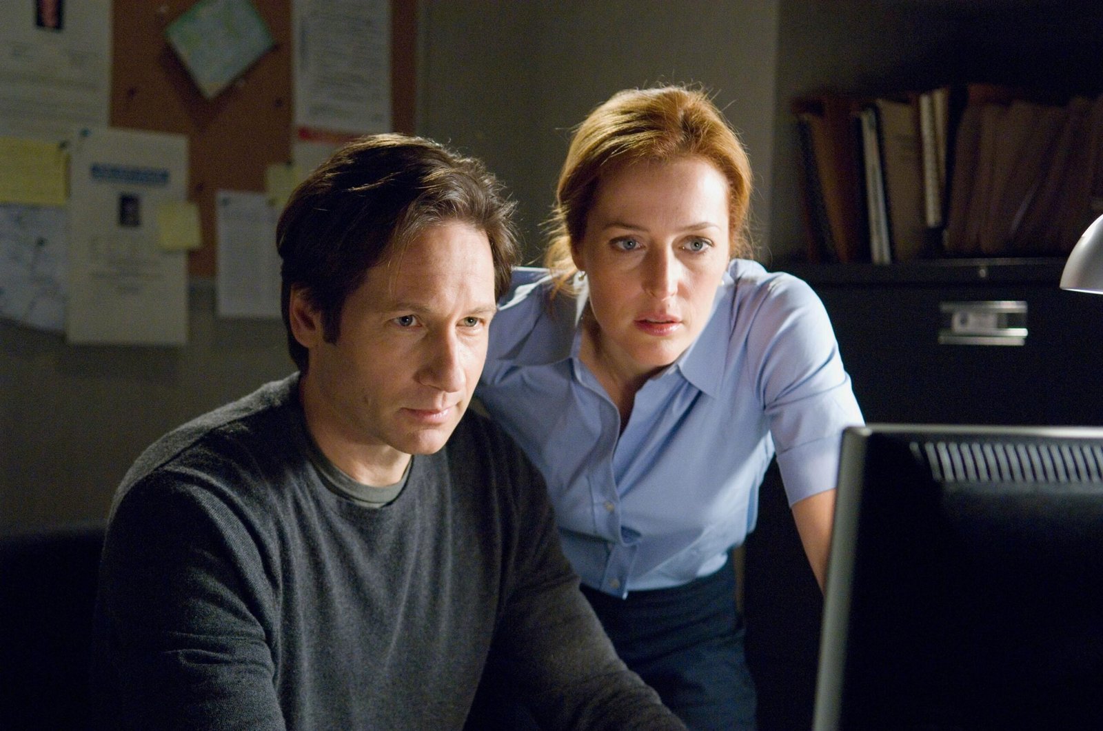 A man sitting at a desk and a woman leaning down over the man’s shoulder. Both are looking at a computer screen.