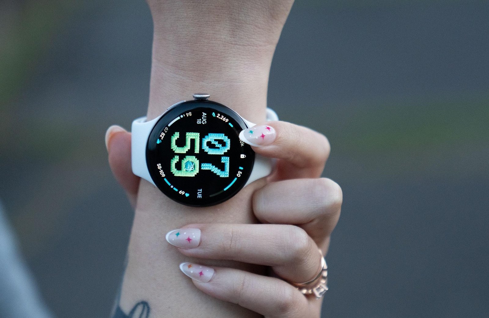 Close up of the Pixel Watch 5 with an AI generated watch face depicting cross-stitch texture on a retro font with some cute and fat woodland creatures in the numbers in a blue and green theme.