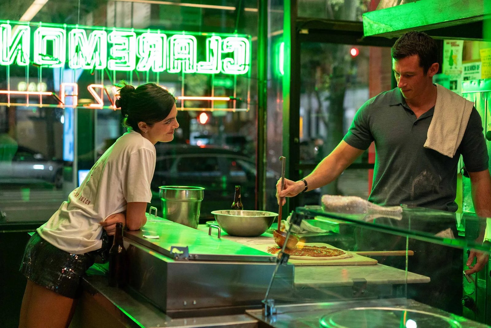 two young people in a pizza shop, he makes pizza, she leans on the counter