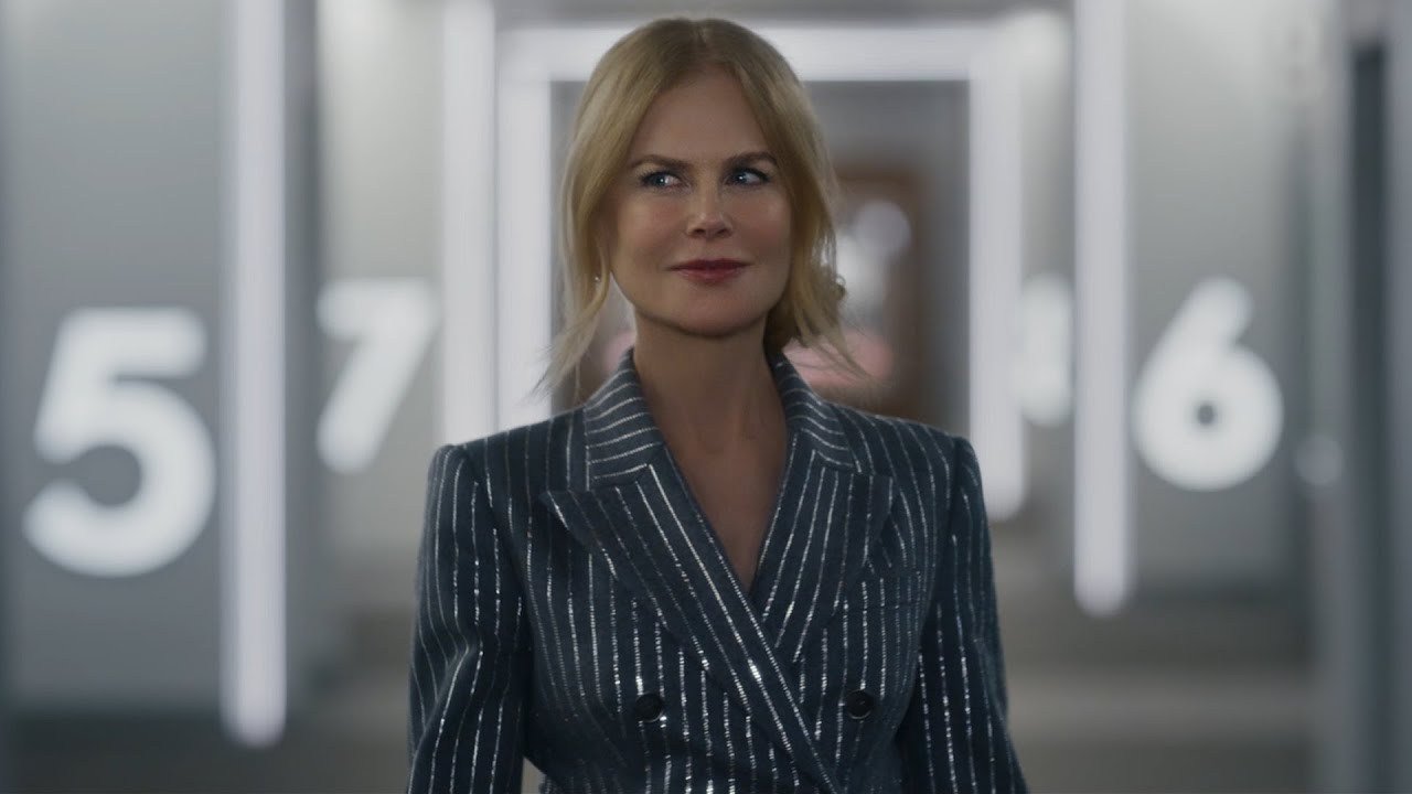 A woman in a glittering suit looking to her right as she walks down a silvery hallway. On both sides, she’s flanked by numbers denoting different movie theater auditoriums.
