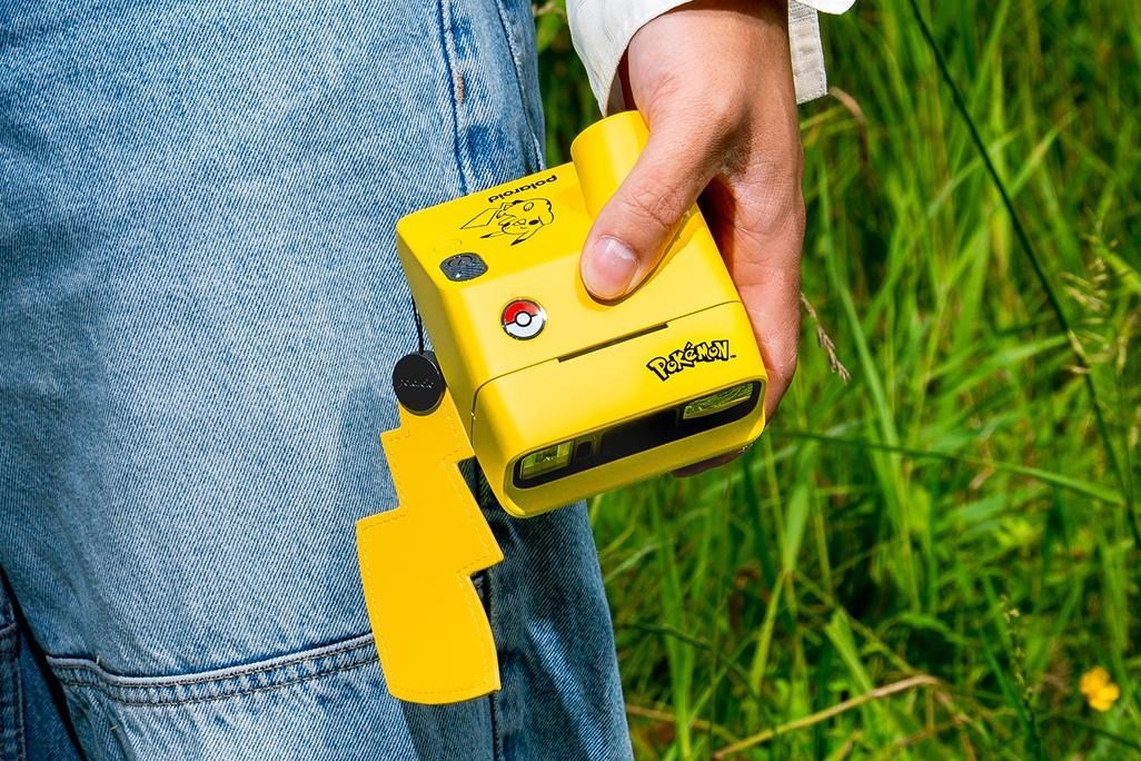 A person standing in grass holds the Polaroid Go Gen 3 instant camera with a bright yellow Pikachu design.